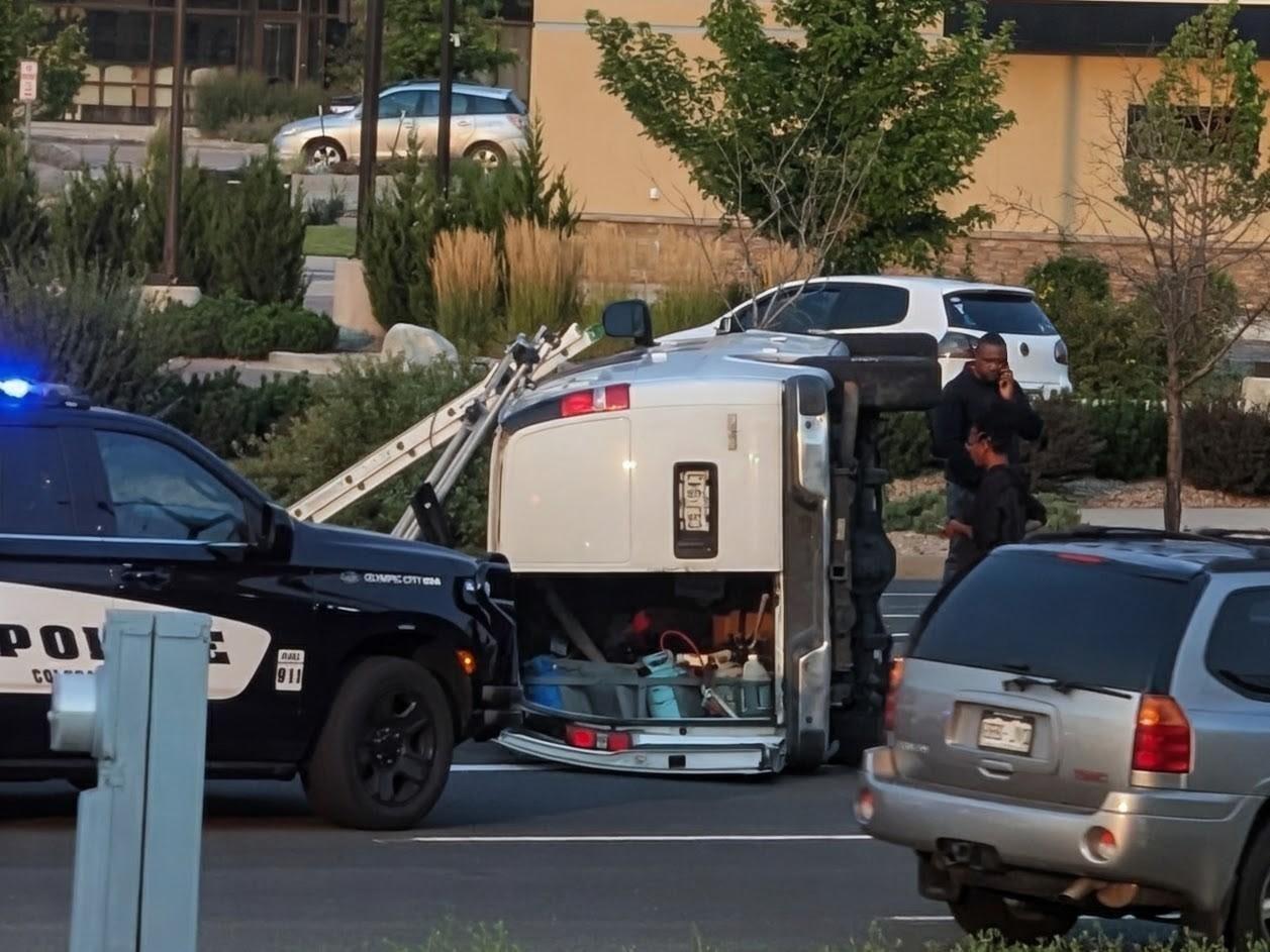 A utility van is lying on its side in the middle of a street, surrounded by police cars and people. The van's side door is open, revealing equipment inside. A police car is parked next to the van, with its lights flashing. In the background, there are other parked cars, some trees, and a building.
