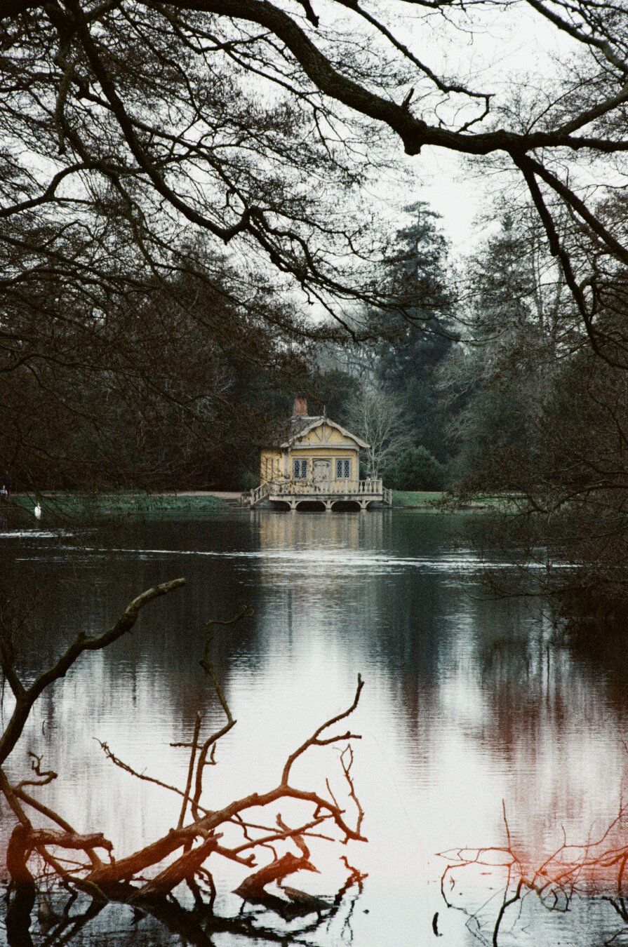 A colour photo of a boat house at the edge of a small lake. The house and some foliage are reflected in the calm waters.
