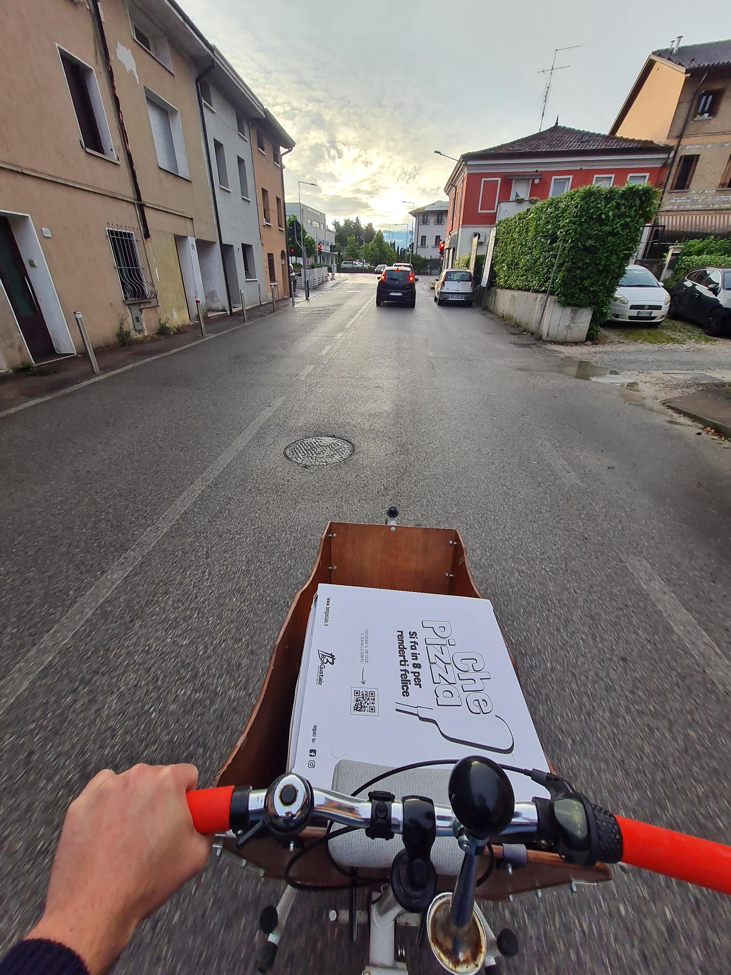Fotografia, presa dal punto di vista del conducente, di una bicicletta cargo in mezzo a una corsia di una strada. Il cassone anteriore della bici trasporta dei cartoni di pizza (maxi, e sono sette).