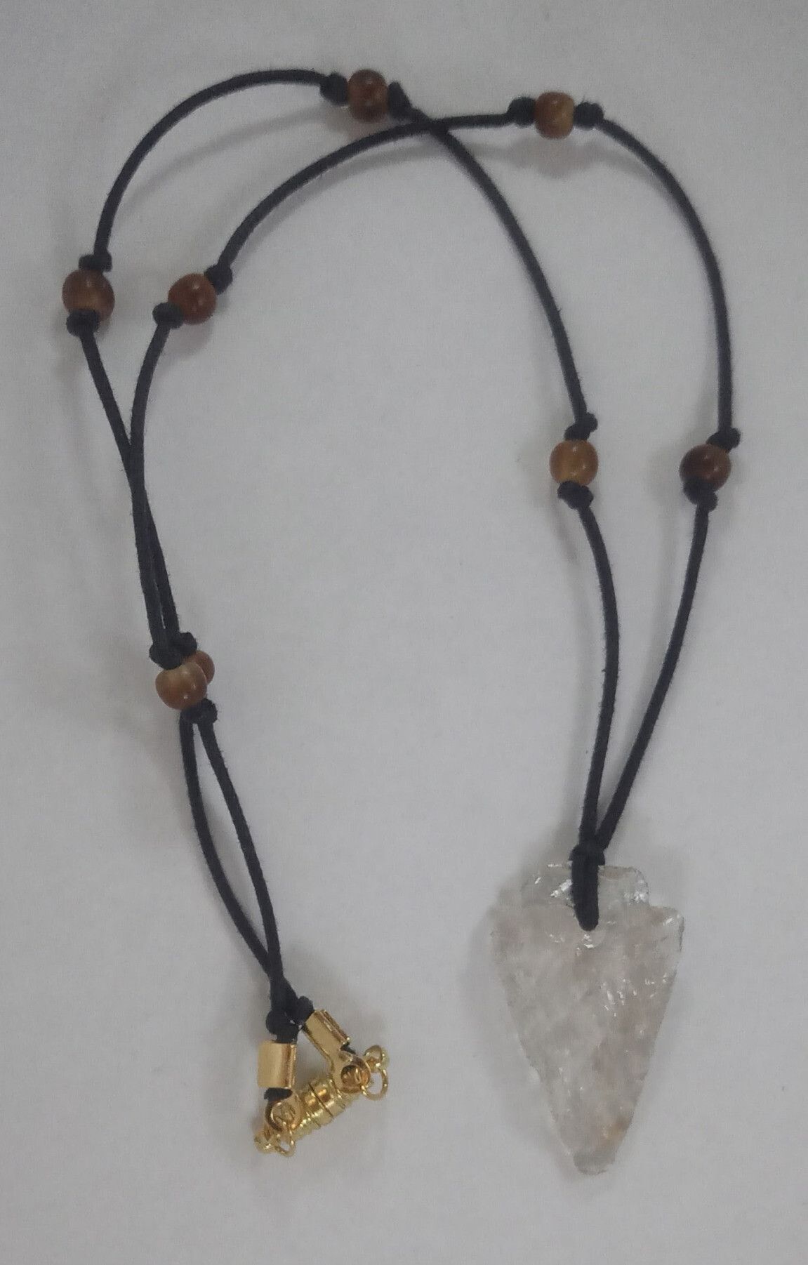 A clear quartz arrowhead pendant, on a white background; the arrowhead has been knapped, so it has lots of rough, chipped-looking edges. It's strung on a black cord, with spherical golden horn beads at short intervals, and a gold-finished oblong magnetic clasp. The cord has been looped through a hole in the top of the arrowhead to hold it. 