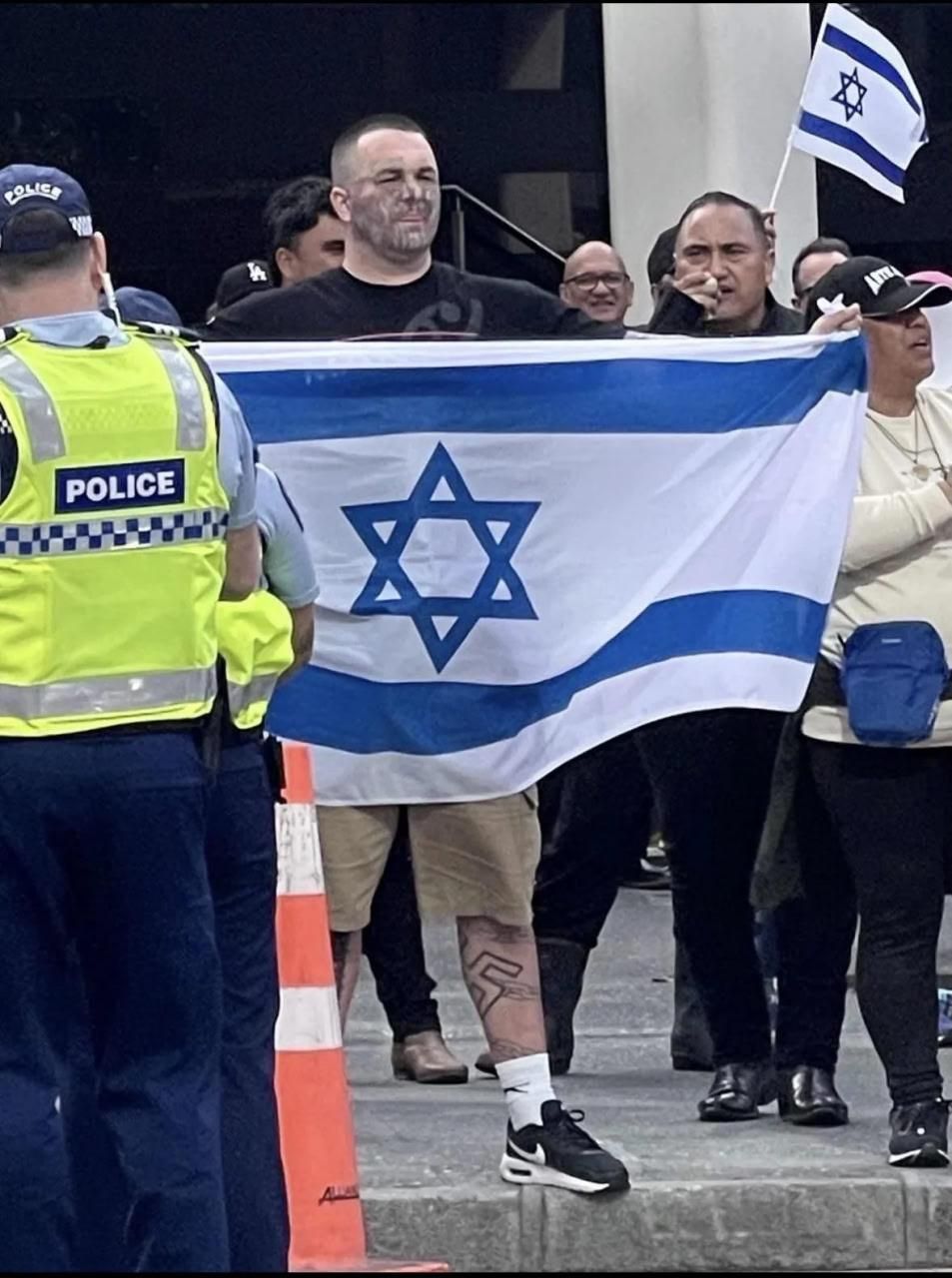 A picture of a protester holding an Israeli flag, based on the police garb visible in the picture this is probably in the UK. The protester has extensive tattos on his face and a gargantuan swastika tattoed on the left calf, half visible in the picture.