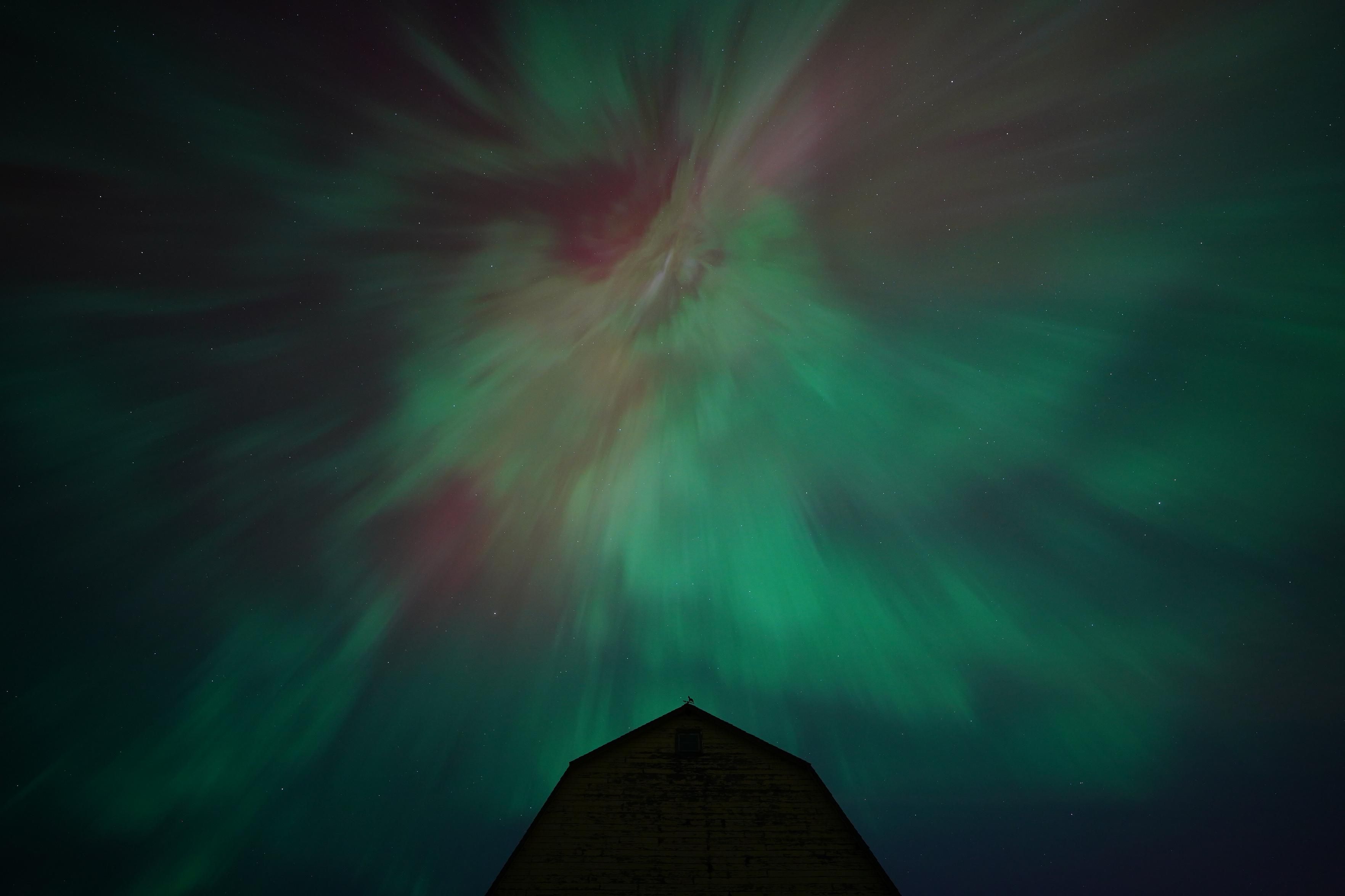 Bright green and red auroras mixed together, but instead of viewing the vertical columns close to the horizon, the vertical columns are directly overhead, so they appear to converge on a point directly above a silhouetted barn.  It looks like something out of a horror film.