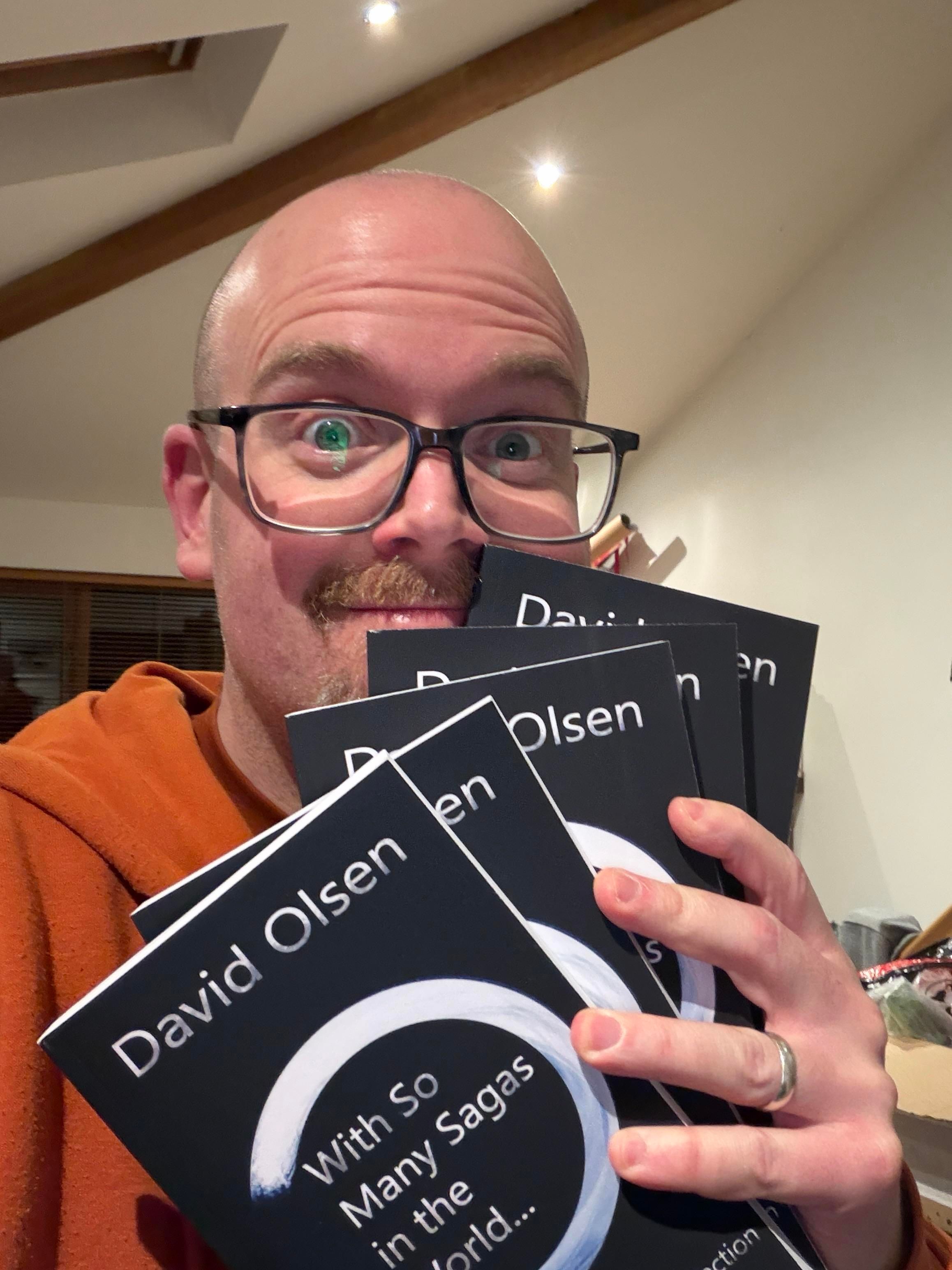 A man with glasses is smiling and holding several copies of a book titled "With So Many Sagas in the World..." by David Olsen. The background shows a cozy room setting with warm lighting.