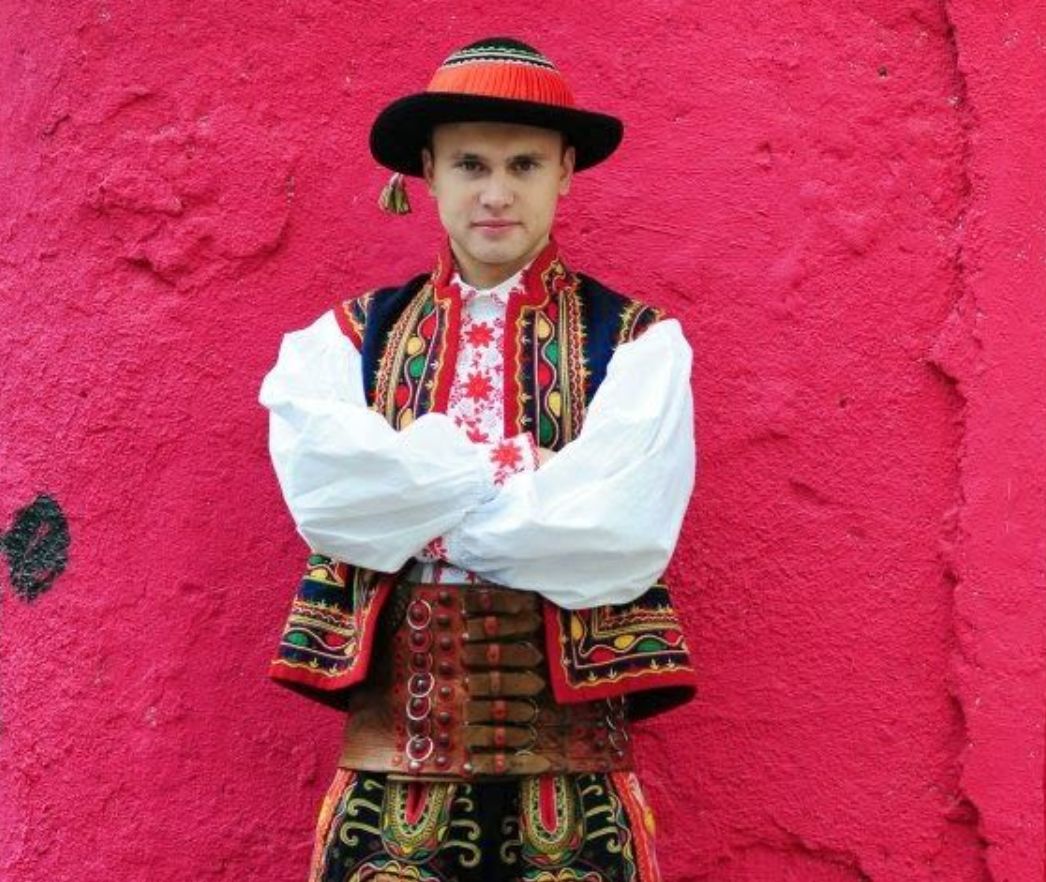 A man stands with arms crossed wearing a traditional highly decorated Polish costume.