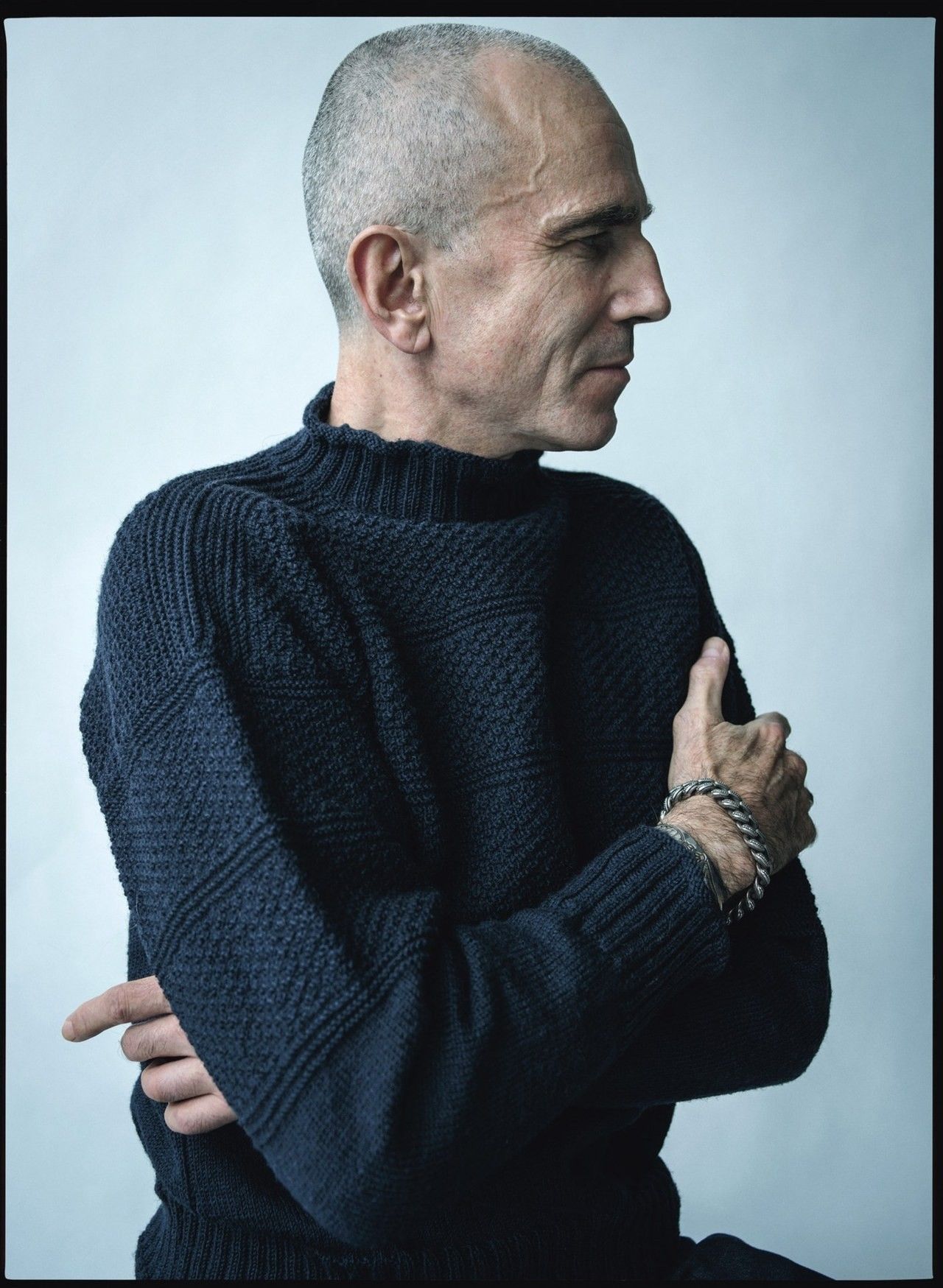Actor Daniel Day-Lewish wearing a navy blue fisherman's gansey. He has his arms crossed over himself. There are three bands of moss stitch separated by alternating rows of knit and purl stitches, and two band of the same moss stitch on the upper arms. There is a slight flare to the mock neck collar, which has been knitted in 2x2 rib, as have the sleeve cuffs.