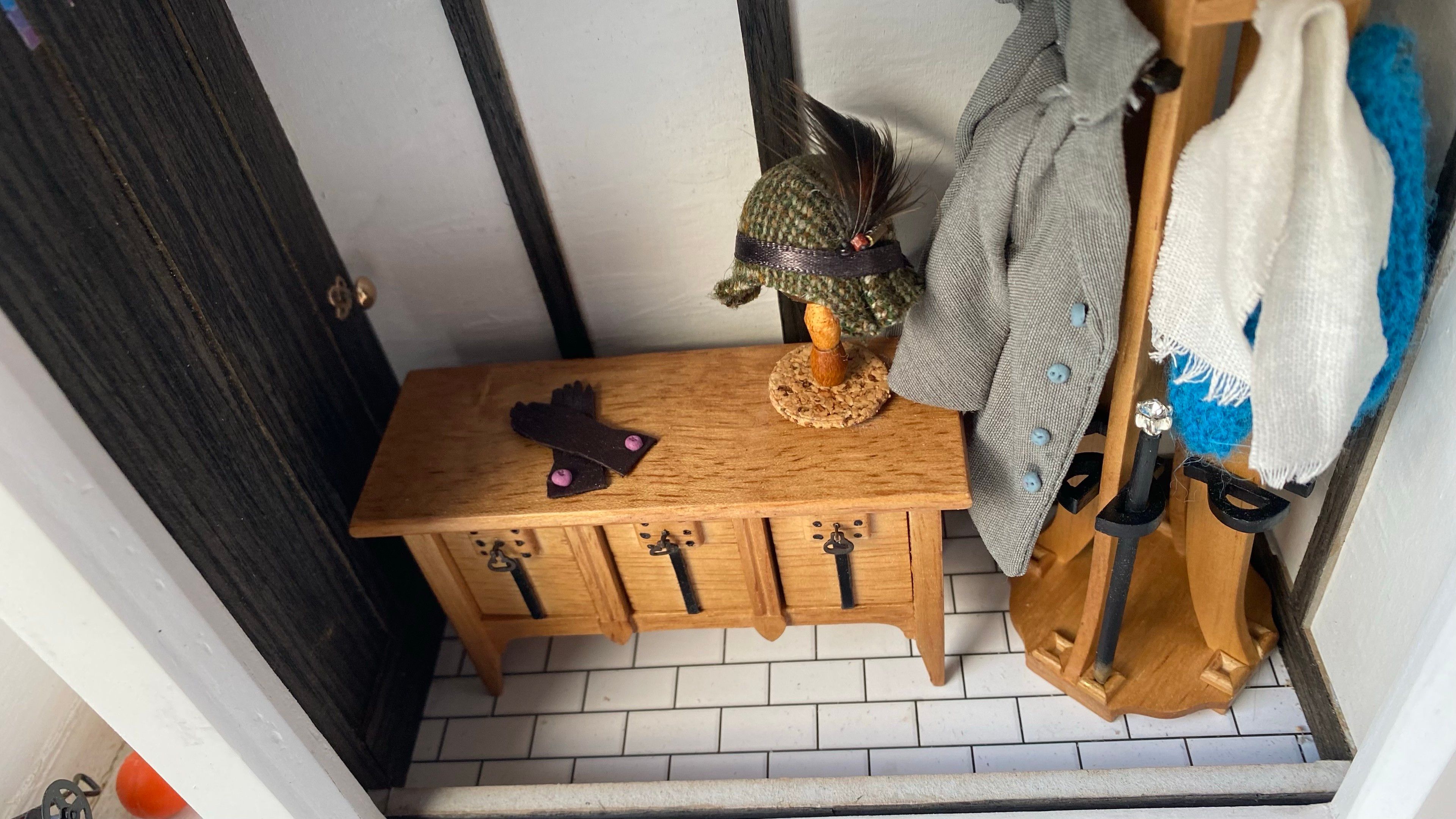 The same miniature hallway scene from above, with a wooden bench, a hat on a stand, gloves, a walking cane, a coat white scarf and blue shawl.