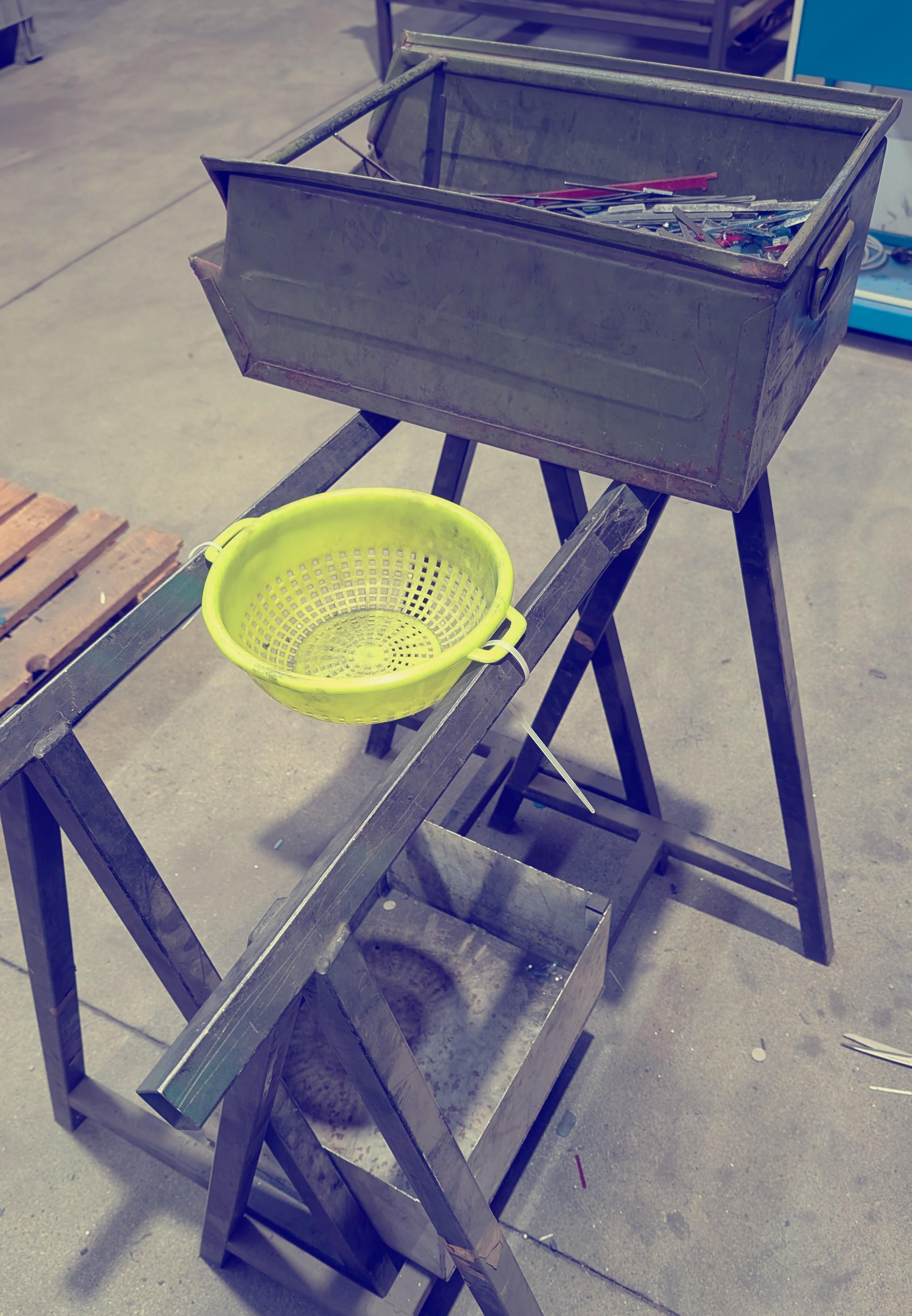 Uno scolapasta verde di plastica è appoggiato su due trespoli ed è tenuto fermo con fascette di plastica. Sotto lo scolapasta un contenitore in ferro per raccogliere gli scarti e sullo sfondo il contenitore da passare al setaccio.