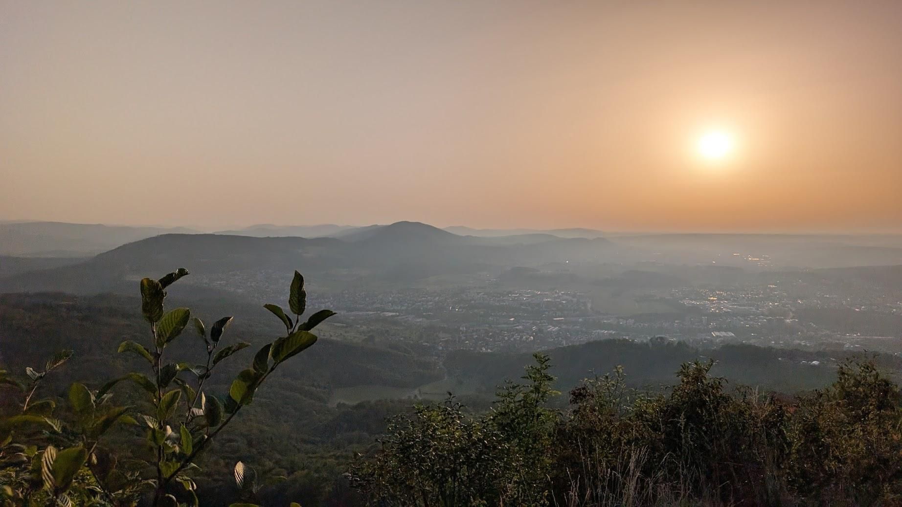 Photo of hills in the distance, close to sunset. The Sun is still a little up. There is a twig in the foreground. The sky is red-ish.