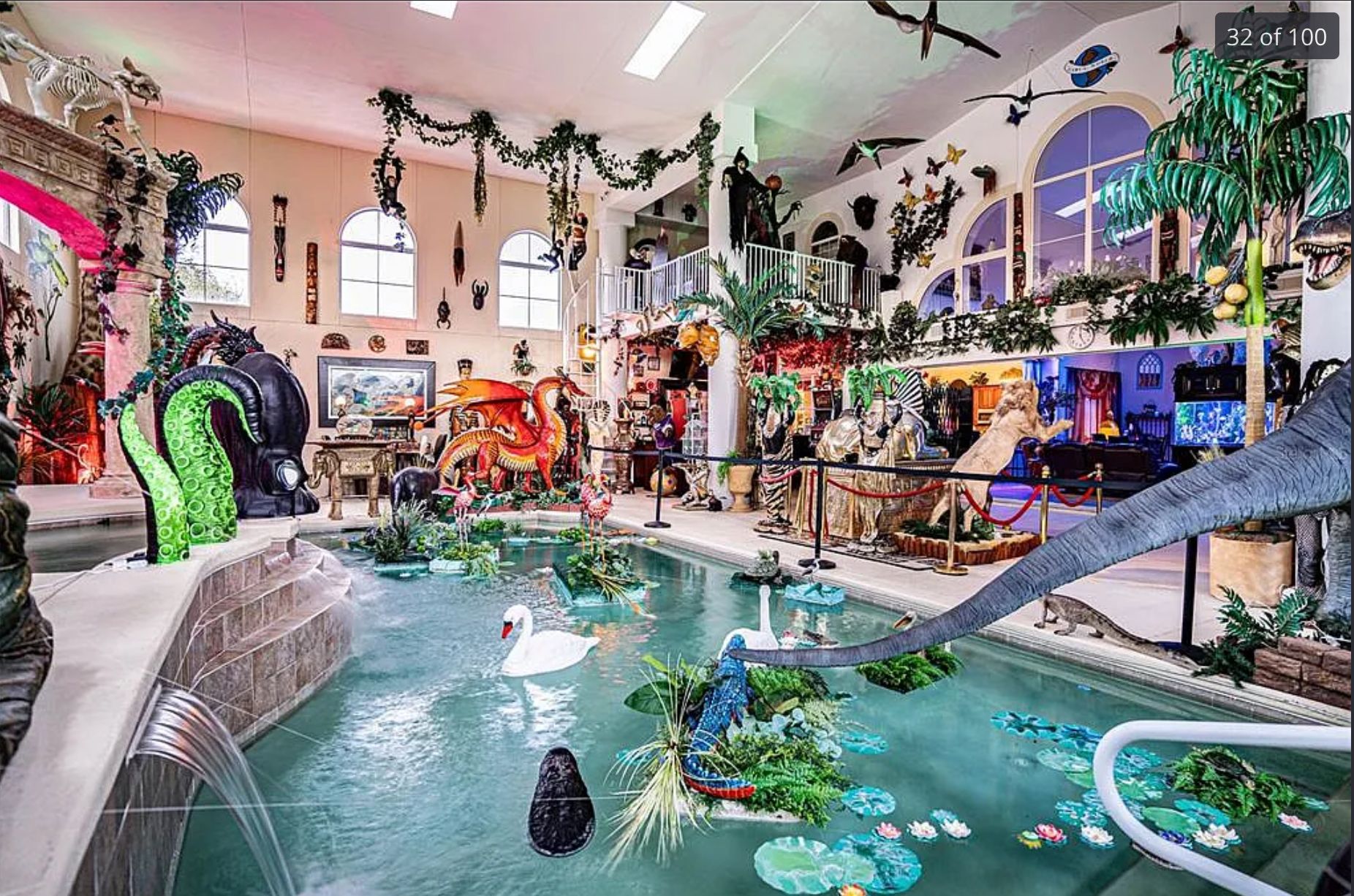 The interior centre of a house which consists of a large pool and an absolute slew of the most random stuff including fake swans, tentacles, dinosaurs, fake birds on the wall, a cat skeleton on top of a fake marble archway, a dragon, a robot, and a fake palm tree. 