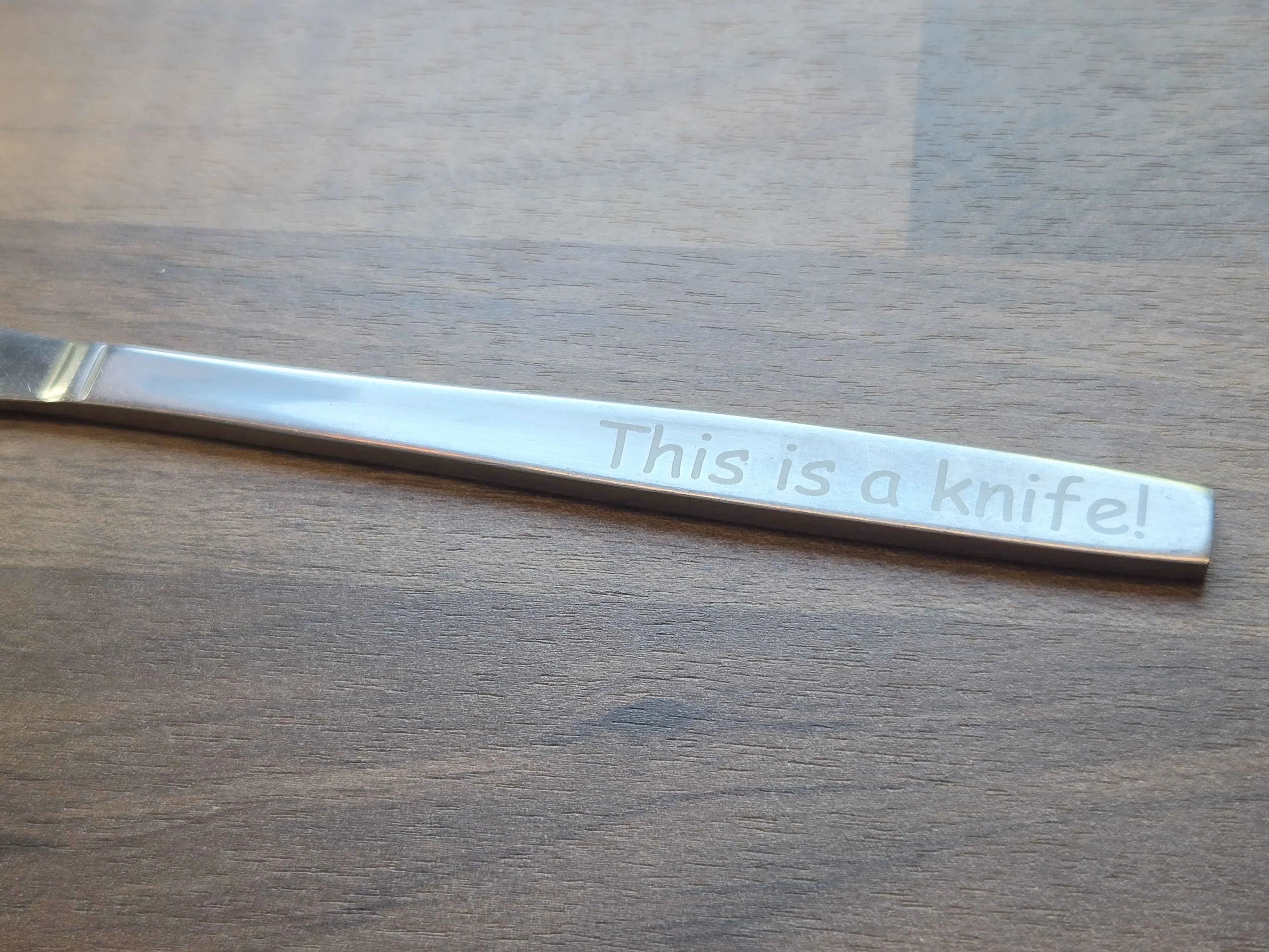 Handle of a metal knife with the phrase "This is a knife!" engraved on it in comic sans.