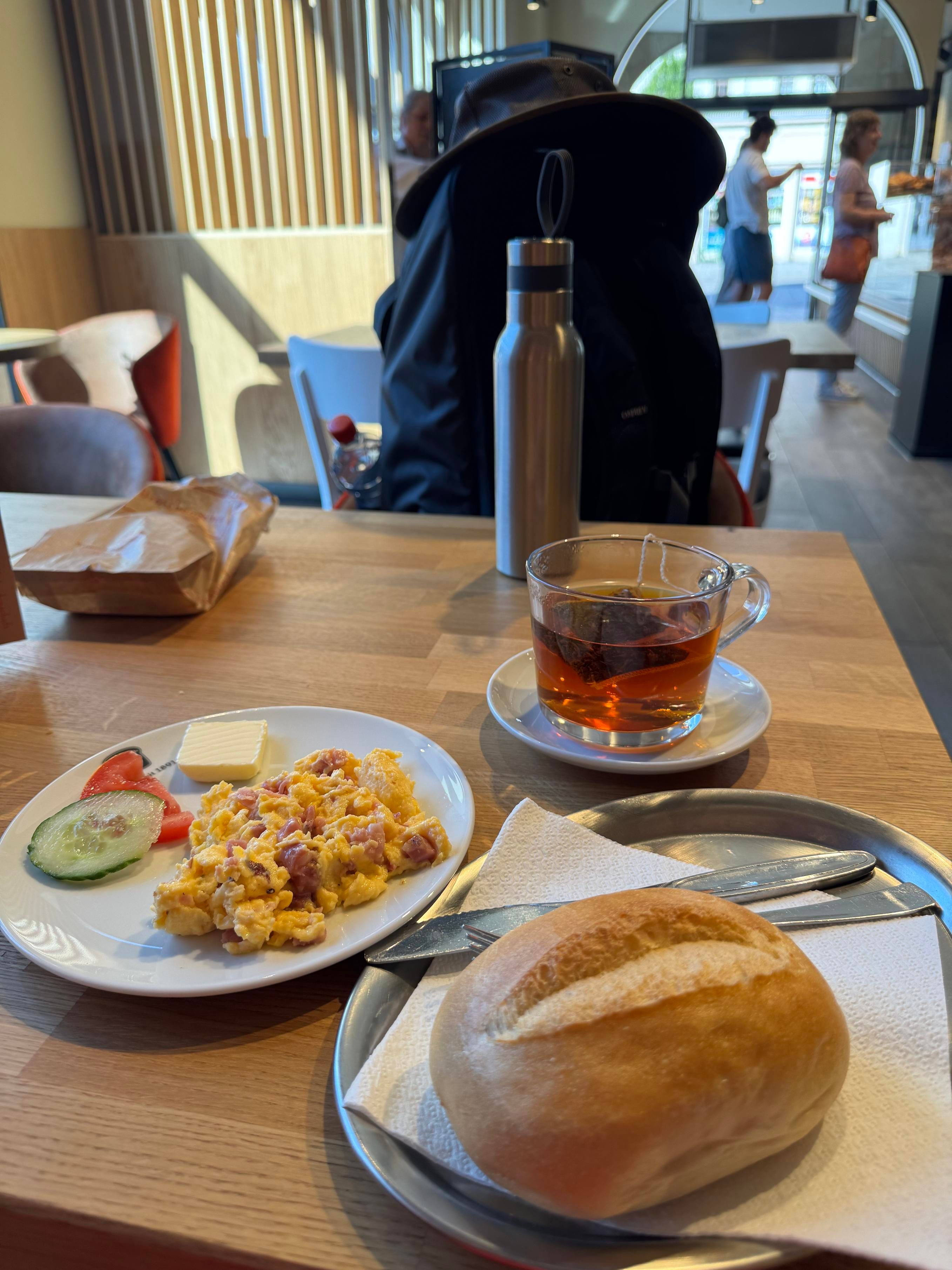 A breakfast consisting of scrambled eggs with ham, butter, a slice of cucumber and tomato, a bread roll and some tea. Behind it is a metal water bottle, and behind that is my backpack topped with my hat