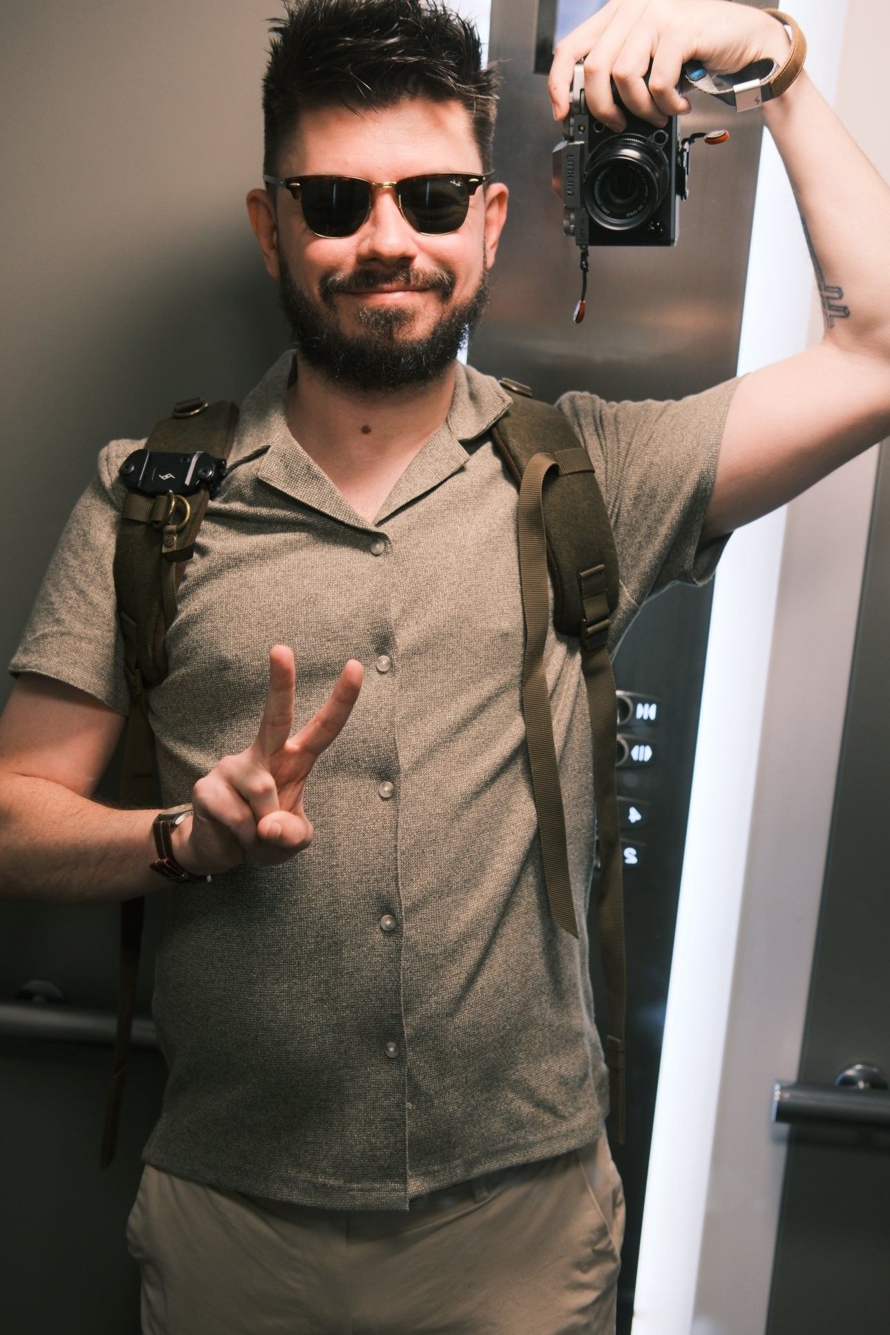 A picture of me in my lift mirror.  I'm holding my camera with a 35mm lens at head height and slightly behind my head to get the framing, which I still miss a bit as my hair and an elbow are slightly cut out.

The picture has warm temperature and enhanced contrast.  I'm wearing a dark green/brown Cuban collar shirt with chinos.  I have a rucksack.   I have short, messy, spiked hair and a light beard.  I'm smiling and throwing up a V sign.  I'm wearing RayBan Clubmaster sunglasses.