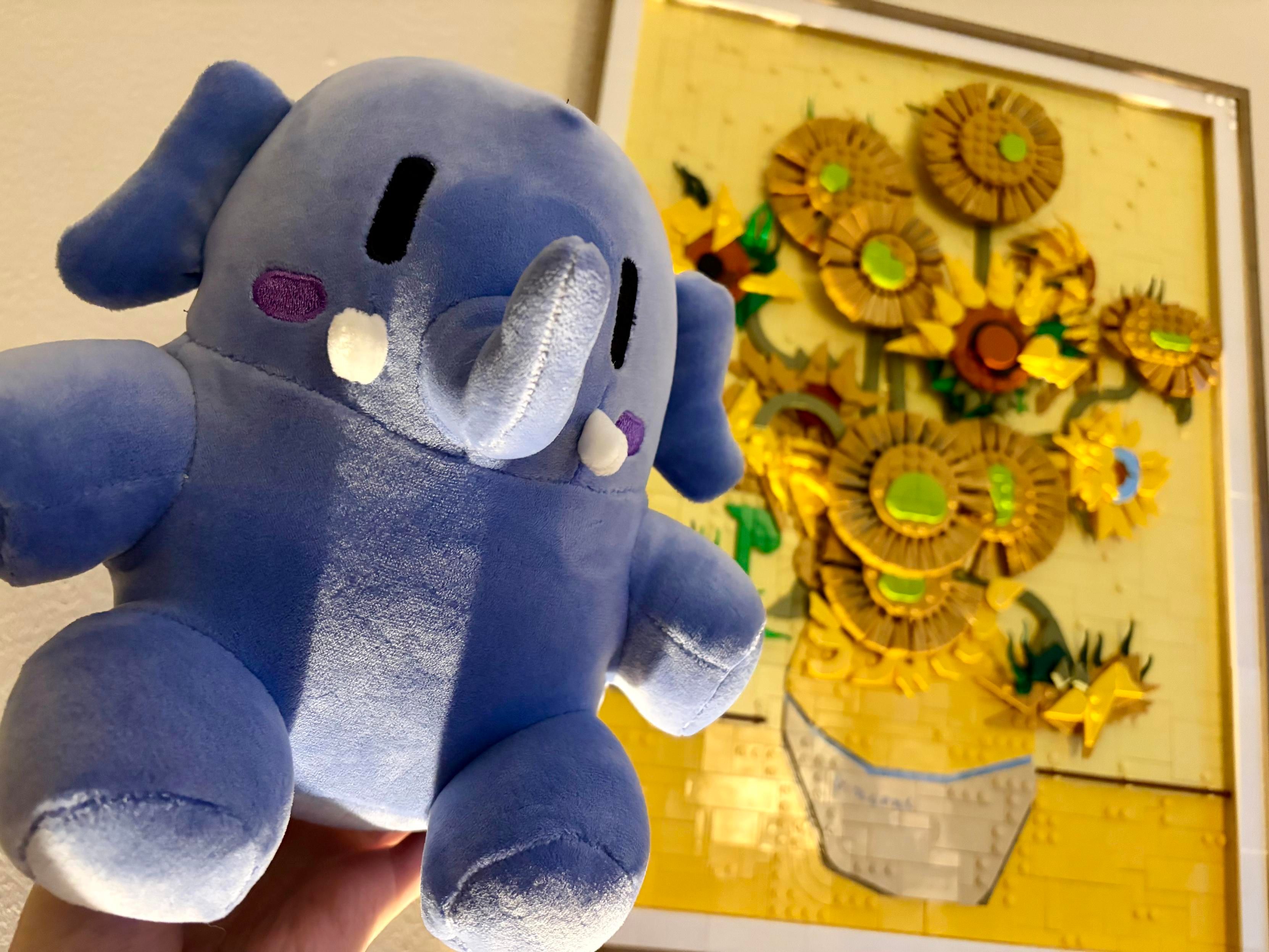 in focus is a dark blue Mastodon plushie, or stuffed animal, that i'm holding up. light is shooting up from below the frame, casting interesting shadows on the plushie. in the background is a bright yellow Lego recreation of a famous Van Gogh painting Sunflowers