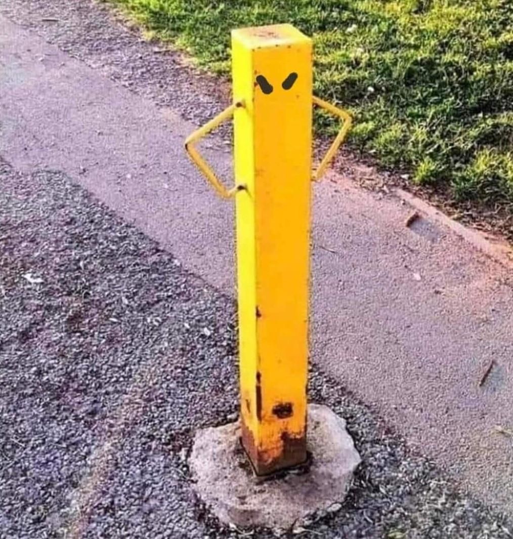 A small yellow bollard with triangles where ropes/chains can be attached that look like little arms akimbo. Someone has drawn on two black lines like frowning eyebrows to complete the indignant look