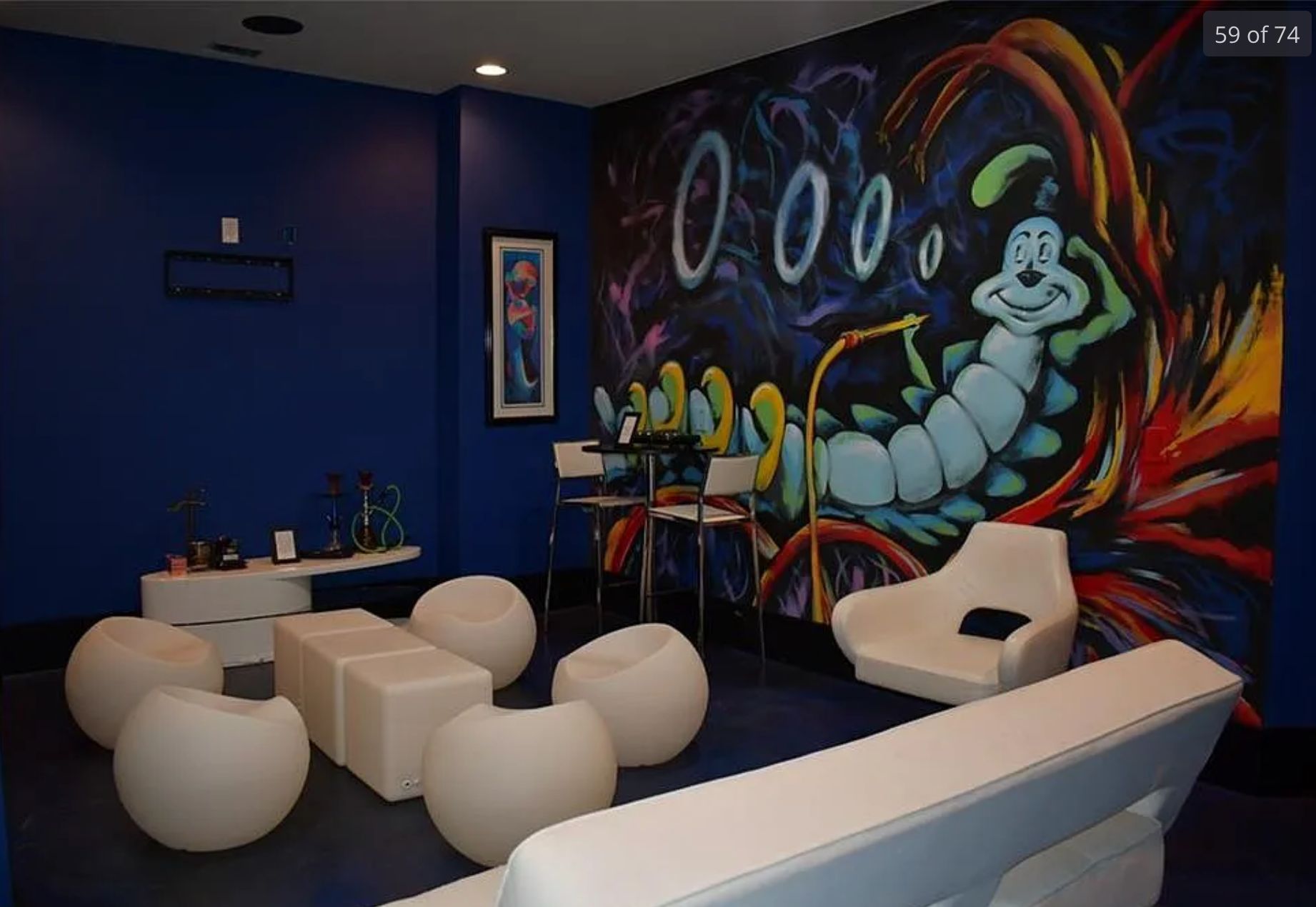 A room with purple walls, white chairs, a hookah, and a wall painted with the blue caterpillar from Alice's Adventures in Wonderland blowing smoke rings from a hookah surrounded by psychedelic swirls. 