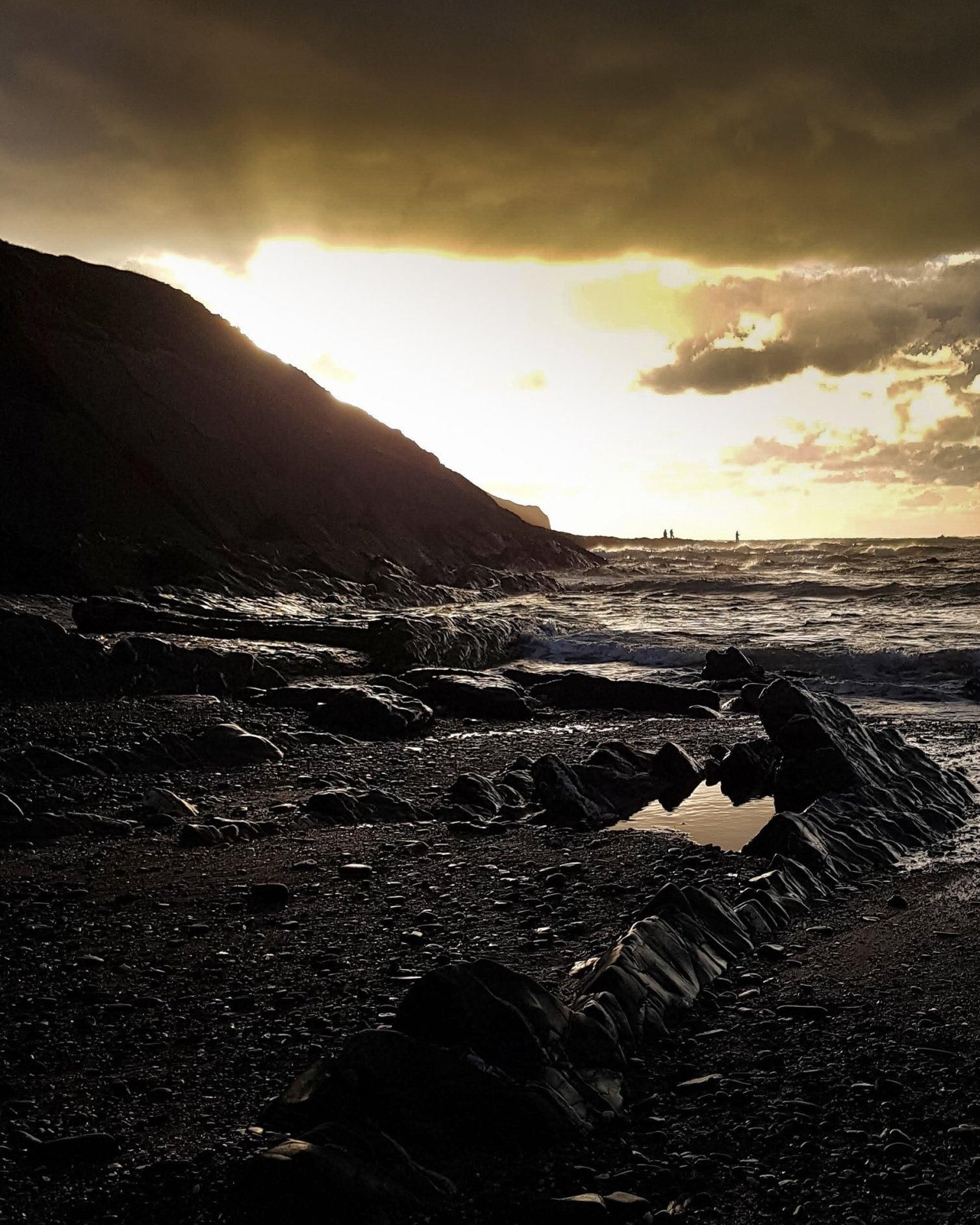 A line of dark, jagged rocks, looking like the fossil of the spines of dinosaur bones sticking out from the shingle beach, jutting out towards the rolling sea. They are in dark shadows cast by a headland immediately to the left of the frame, but picking up the light on one side. A clouded sky above is dimmed by the stormy grey cloud, but lit from underneath by a clearer patch of golden light from the sunset behind the headland. Far out on the horizon, three small figures can be seen, standing on unseen rocks, looking like they're standing in the rolling ocean as they fish.