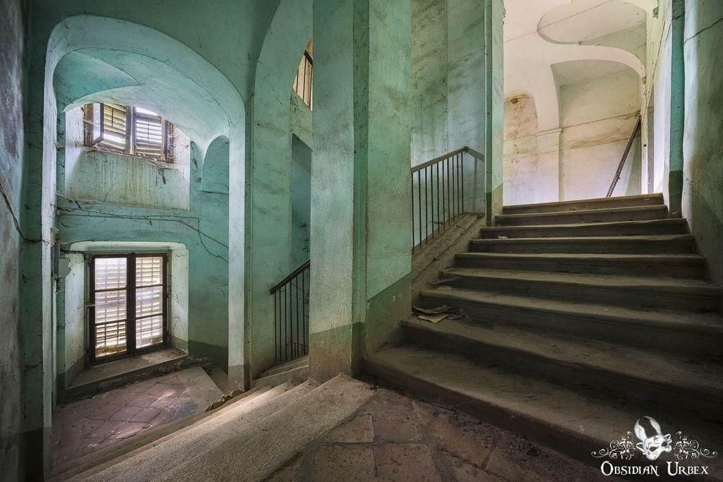 A vintage staircase in a weathered building showcases a unique architectural design. Light streams in from windows, highlighting the textured walls, arches