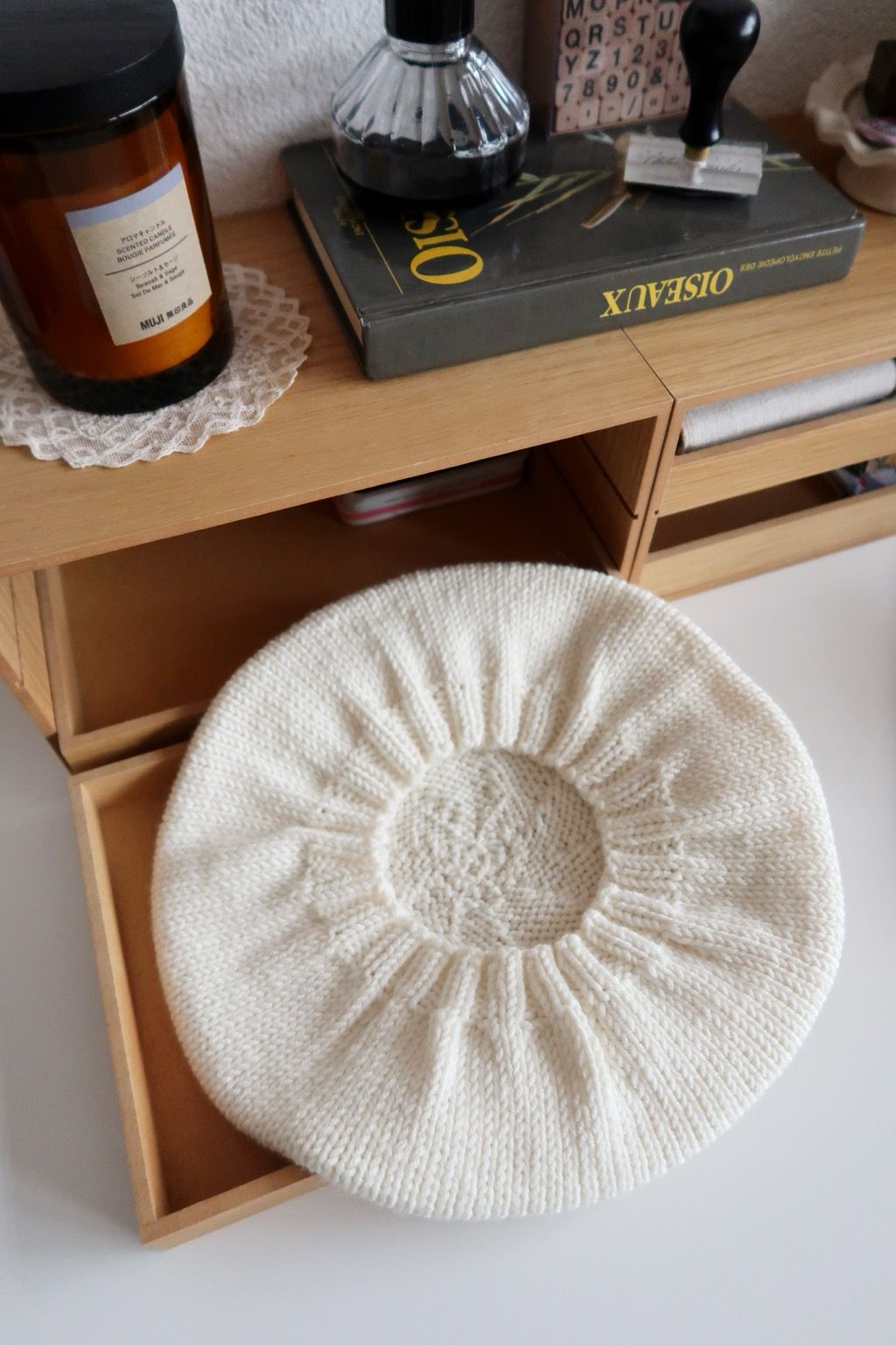 Un béret en tricot vu d’en dessus, de forme ronde avec des détails en spirales, posé sur une petite étagère en bois dont le tiroir est ouvert. Autour, dans le décor, il y a une bougie posée sur un napperon en dentelle, un livre d’ornithologie ainsi qu’un pot d’encre noir, un tampon encreur et un kit alphabétique de mini tampons encreurs.