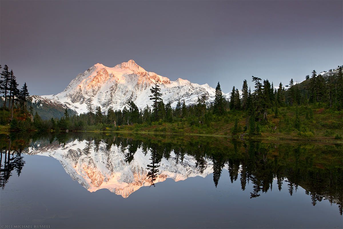 Mount Shuksan's peak reflecting in picture lake during a fall day with a blue sky and a trace of snow around the trees at the edges of the lake.