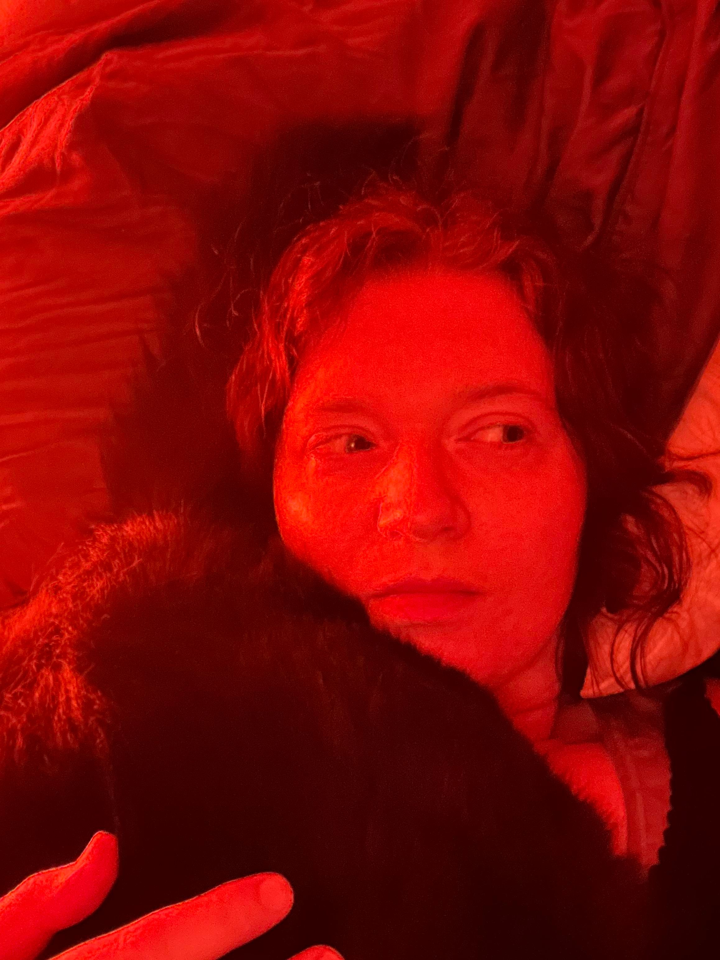 Selfie of my face and shoulders as I'm lying in bed. A fluffy cat backside is lying on my chest and neck and the tail is curled around the side of my head. I'm looking to the side to avoid eye contact for people who don't care for that. The lighting is red because that's apparently a better colour if you have a migraine tendency