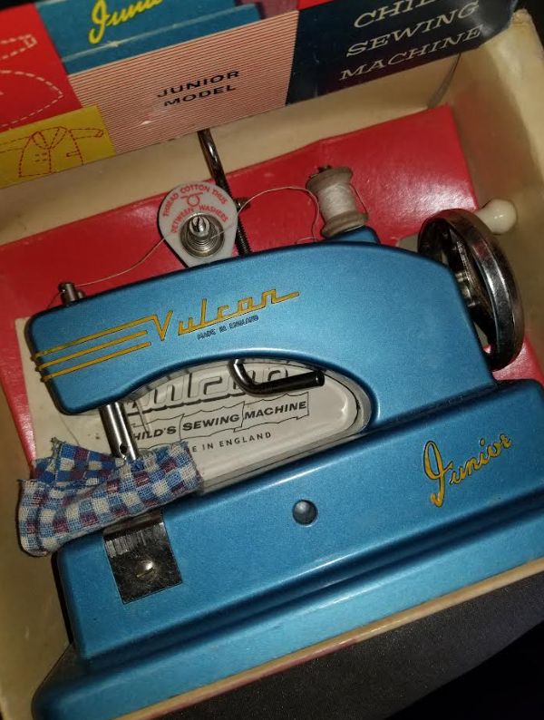 A vintage toy sewing machine lying flat in the box. It is blue. There is some fabric under the presser foot, a blue gingham. It's a little hand crank one, so there's a flywheel on the right and the word Vulcan embossed into the top bit and painted gold. The word Junior is embossed in gold on the base. 