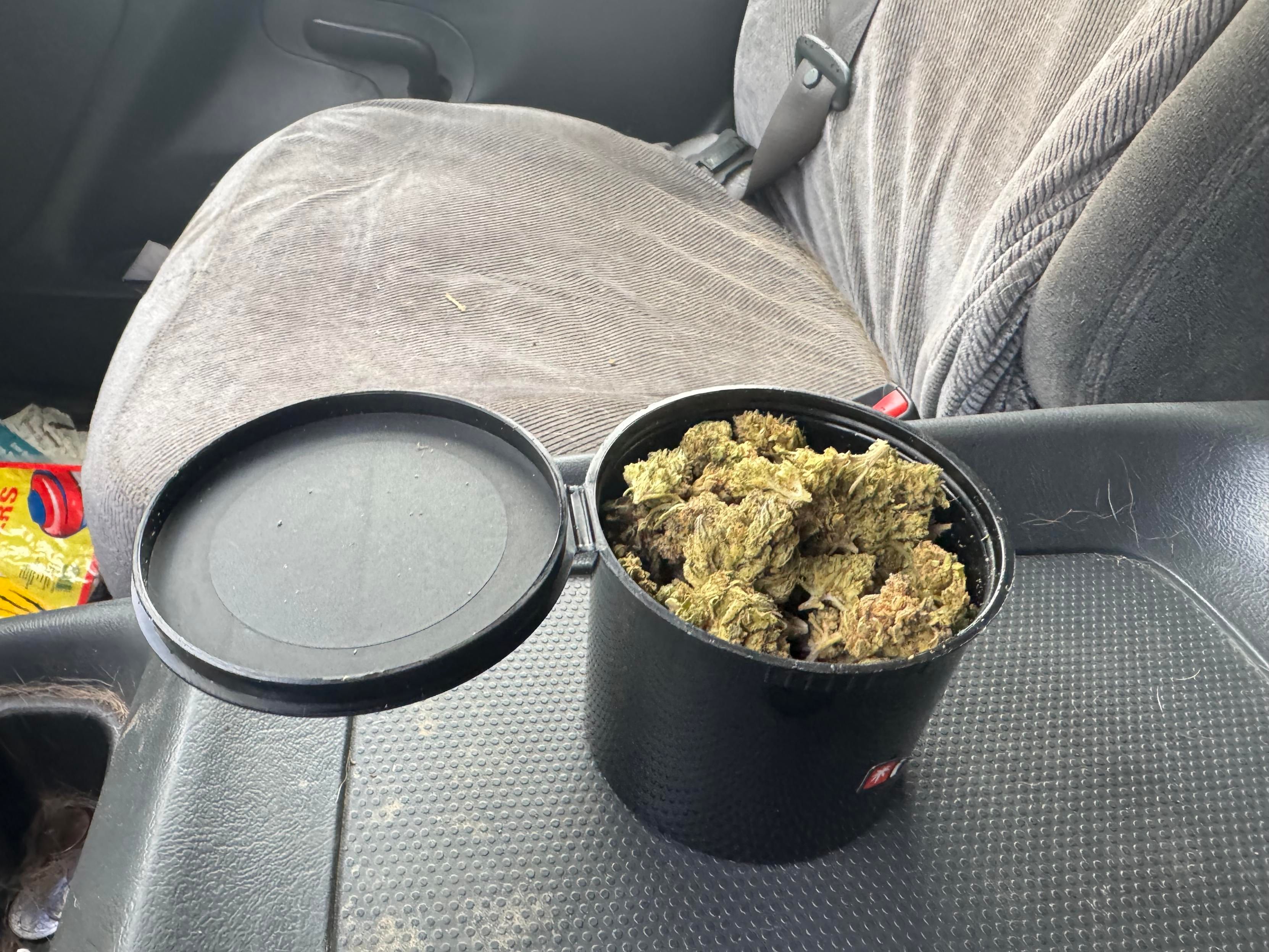 A black container partially open, filled with cannabis buds, is placed on a car seat. The seat is made of fabric and there are items visible in the background.