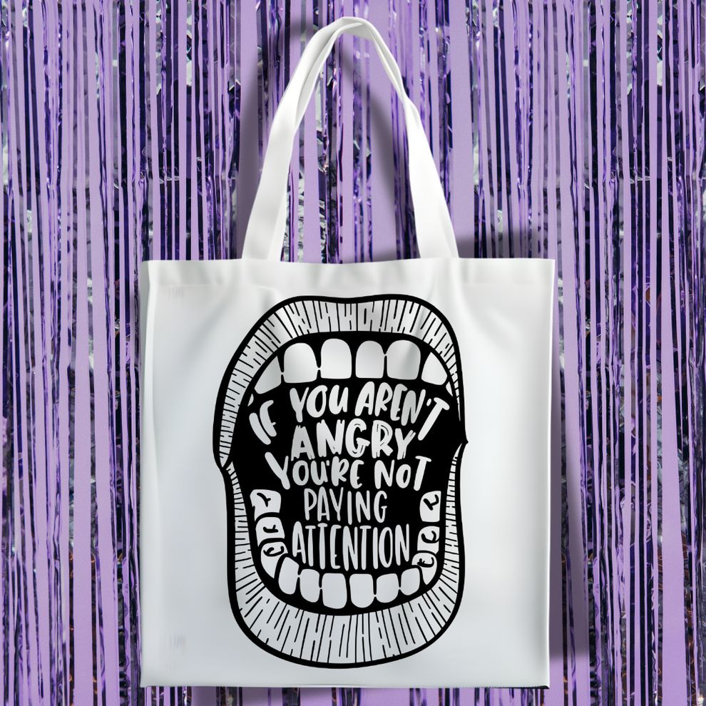A white canvas tote bag featuring a black graphic of an open mouth with teeth. Inside the mouth, the bold text reads, "IF YOU AREN'T ANGRY YOU'RE NOT PAYING ATTENTION." The bag is displayed against a background of shimmering purple tinsel.