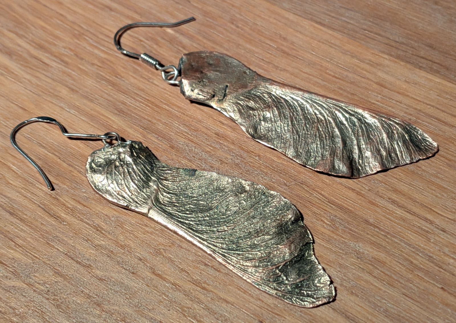 Tilted view of the same two shiny silver maple seeds with earring hooks attached to the top lying on a wooden surface.