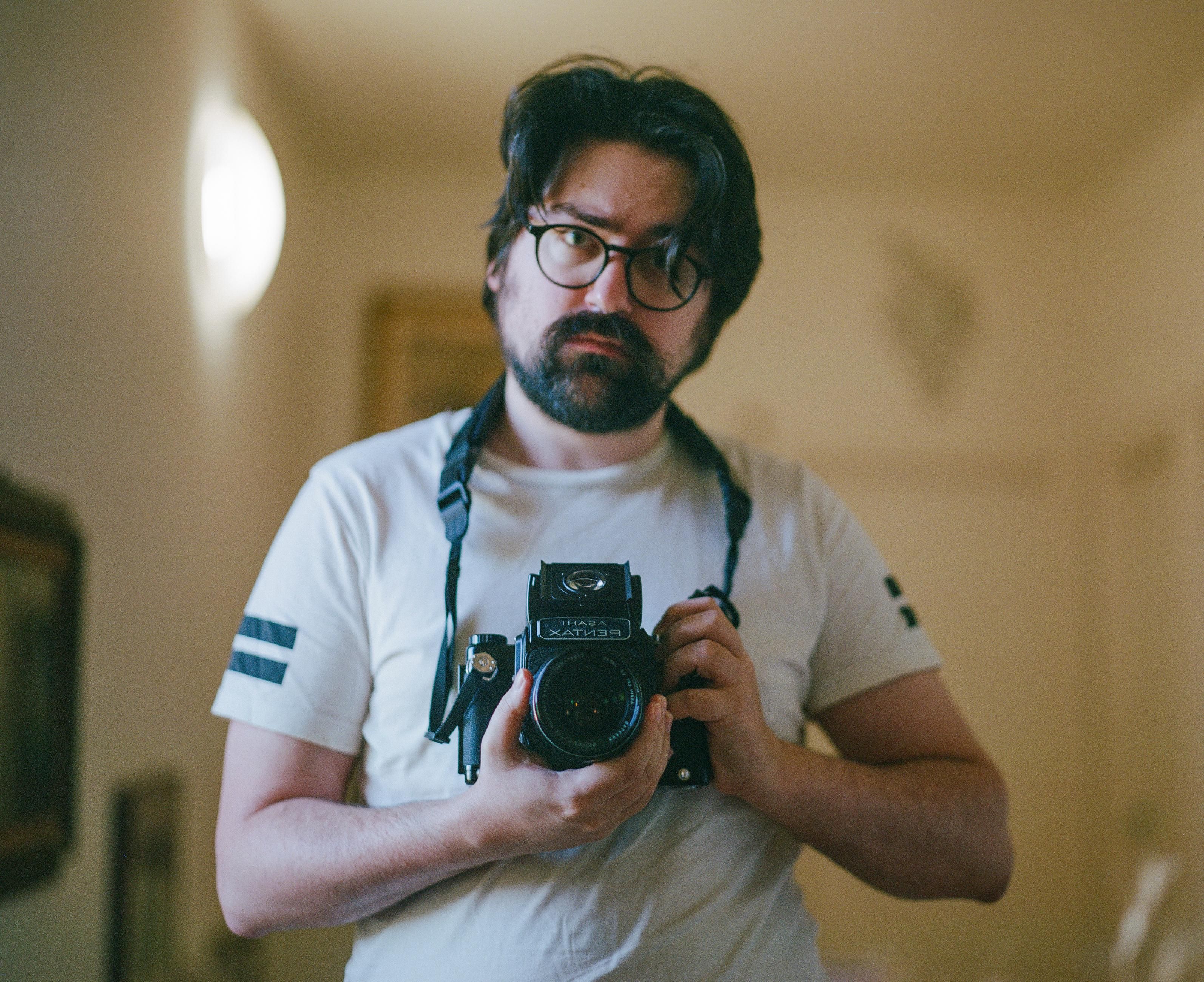 A white man with black medium-length hair and a short beard, wearing round black frame glasses and a white t-shirt, holding a Pentax 6x7 camera with an open waist level viewfinder, looking in the mirror in a big hallway.