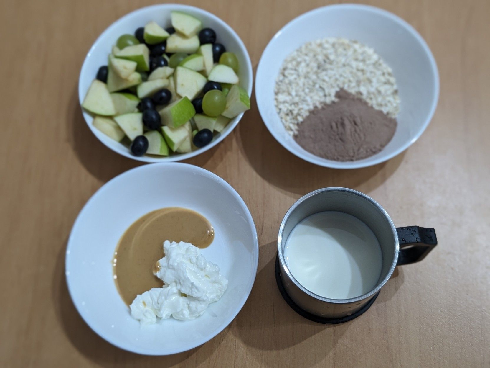 Main ingredients of the milkshake on separate small bowls on a wooden table seen from above:
Bowl 1 on top-left: Fruits (apple, grapes, blueberries)
Bowl 2 on top-right: Outmeal and protein powder
Bowl 3 on bottom-left: Yogurt and peanut butter
Metal cup on bottom-right: Milk