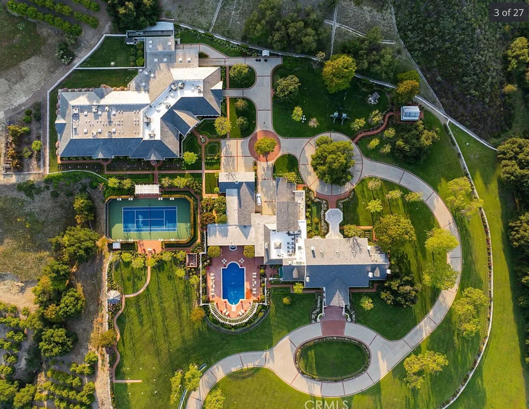 An overhead view of an absolutely immense, sprawling mansion complex with one main house and several other buildings, a pool, tennis court, and landscaping. 