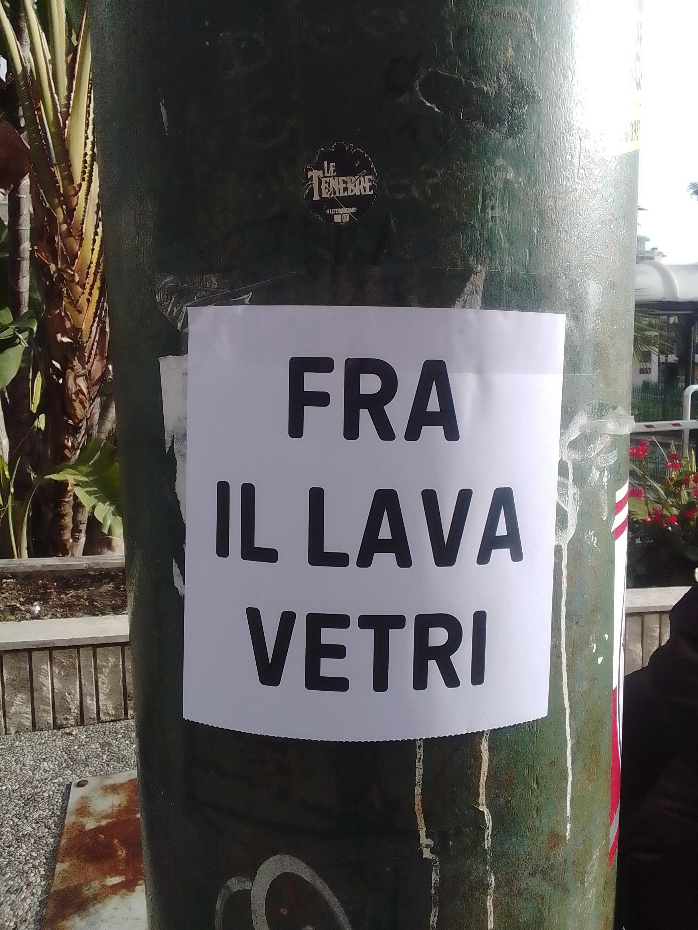 Foto verticale a colori che ritrae un volantino bianco cin una grande scritta in nero, "fra il lava vetri", appiccicato a un grosso palo verde.