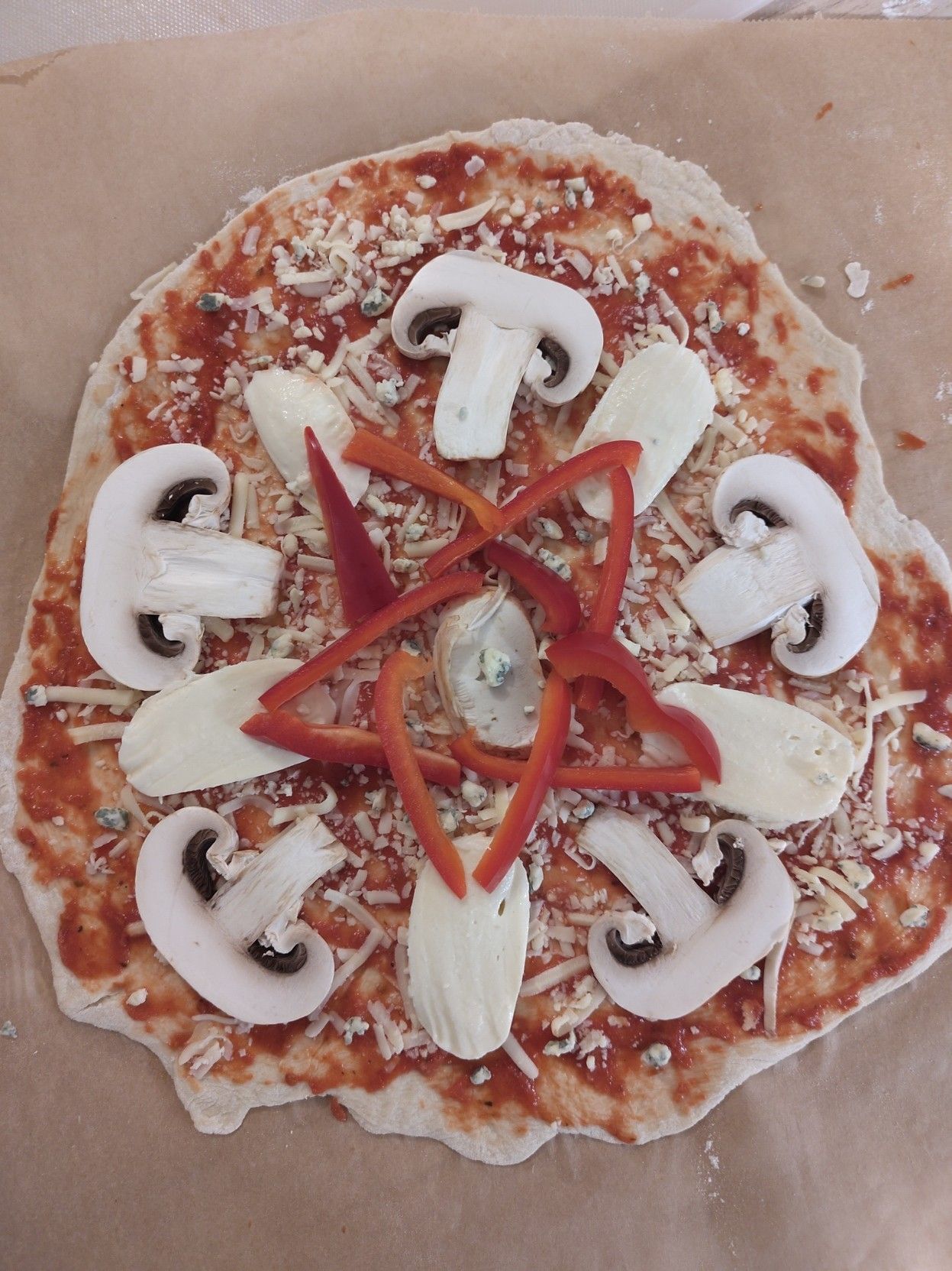 Kuva vielä paistamattomasta pitsasta. Pitsan keskellä on punaisista paprikanpalasista kasattu pentagrammi. Muut täytteet ovat herkkusienen ja mozzarellan viipaleita jotka on aseteltu osoittamaan keskustasta poispäin.