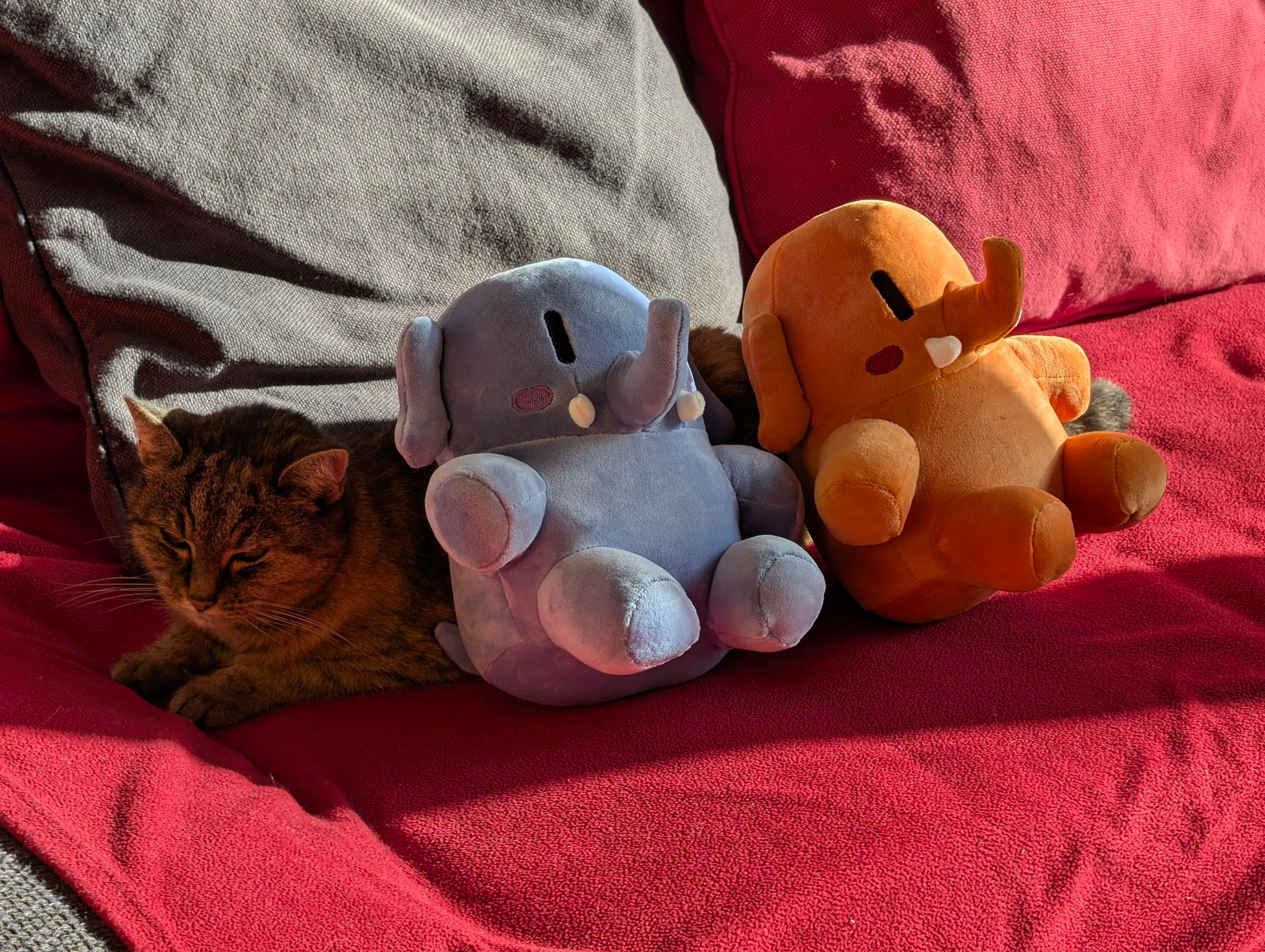 Same as photo 2, but the camera position moved to the left, so the cats face is visible. Now the plushtodons both face away from the cat while sitting leaned back on the side of the cat, as if she was a pillow used as a backrest