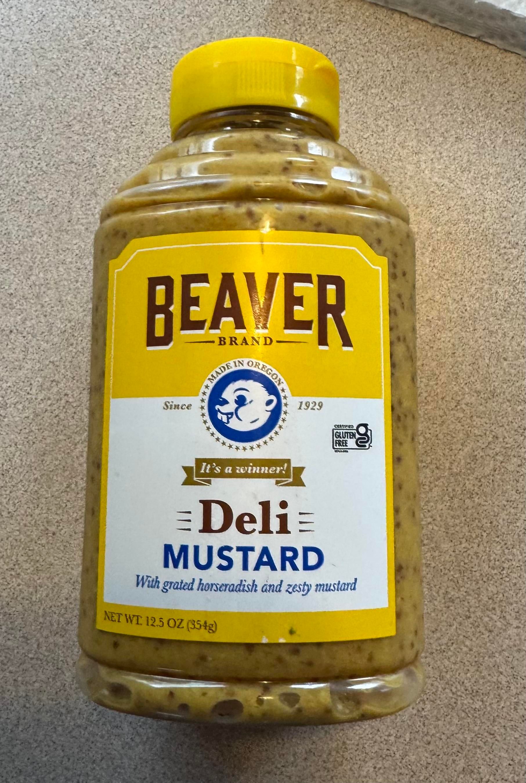 A bottle of Beaver Brand Deli Mustard featuring a yellow cap and prominent labeling. The mustard contains grated horseradish and zesty mustard, and the bottle holds 12.5 ounces. The brand claims to be gluten-free 