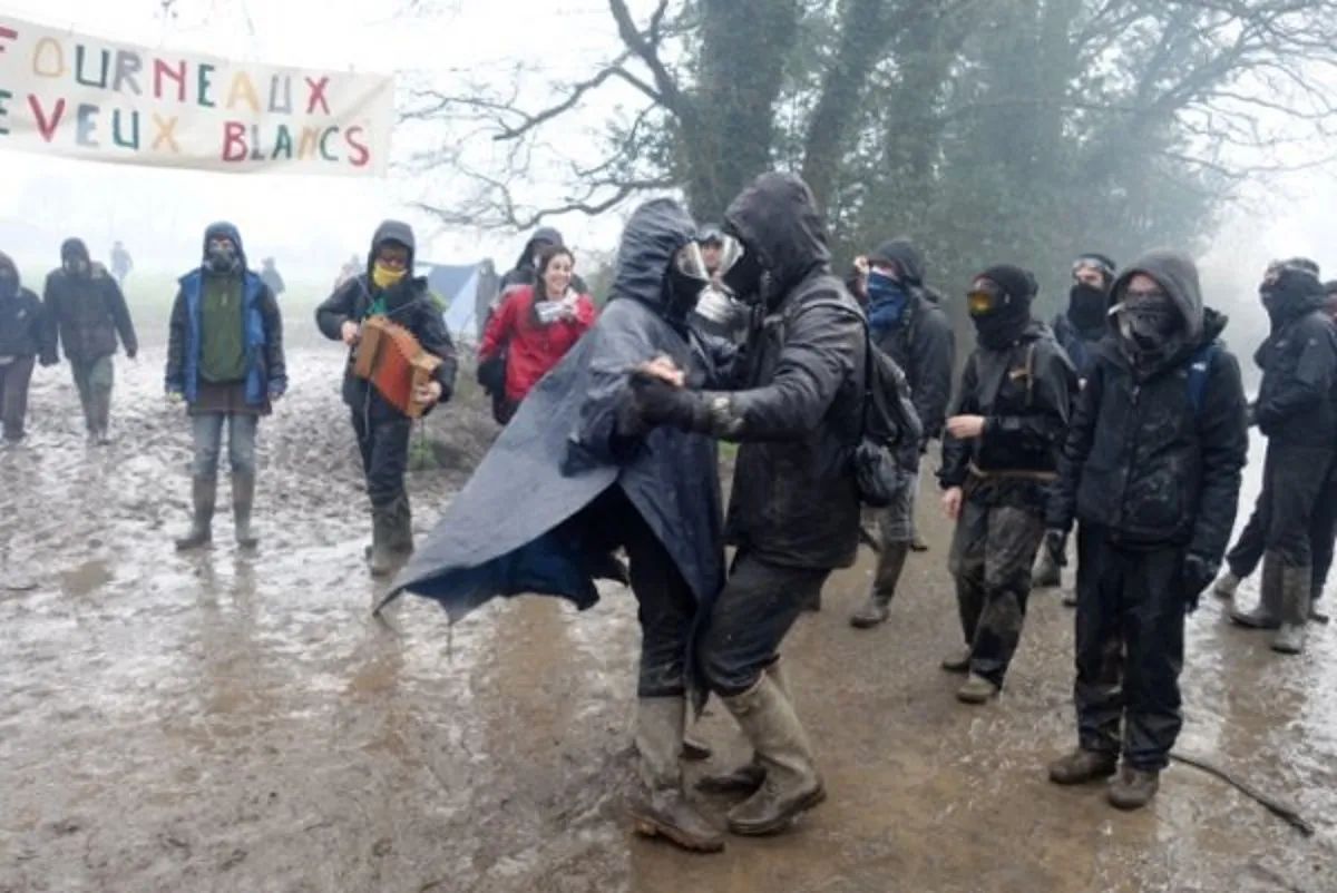 zwei mit Grummistiefeln und schwarzen Regenmänteln sowie Gasmasken, tanzende Menschen im Schlamm. Im Hintergrund ein paar weitere vermummte Menschen. Einer mit einem Akkordeon.
