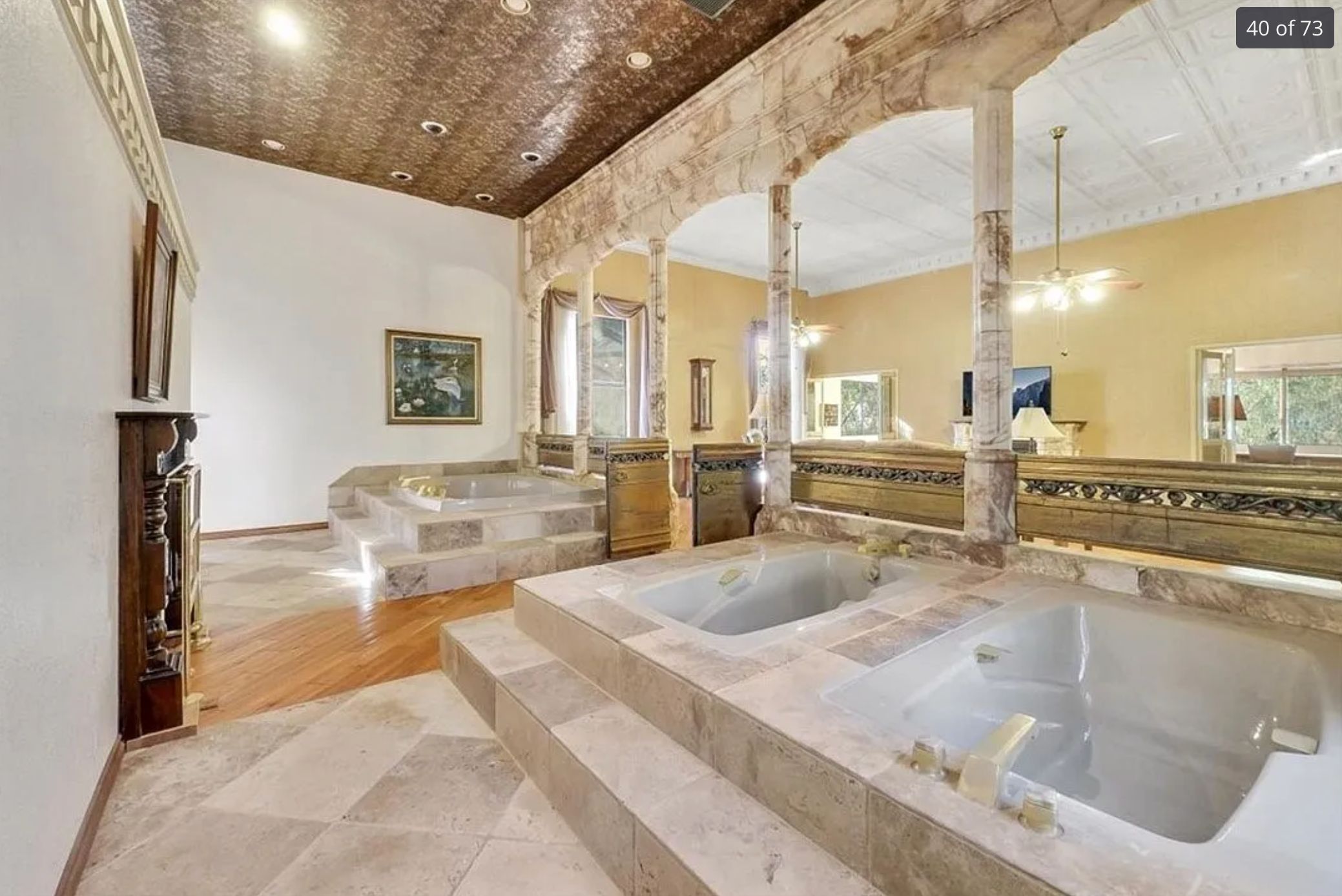 An area with 3 marble jacuzzis in front of a fireplace. 2 are next to each other and another one is slightly further away. The wall behind it has some columns but is otherwise open to the rest of the living area. 