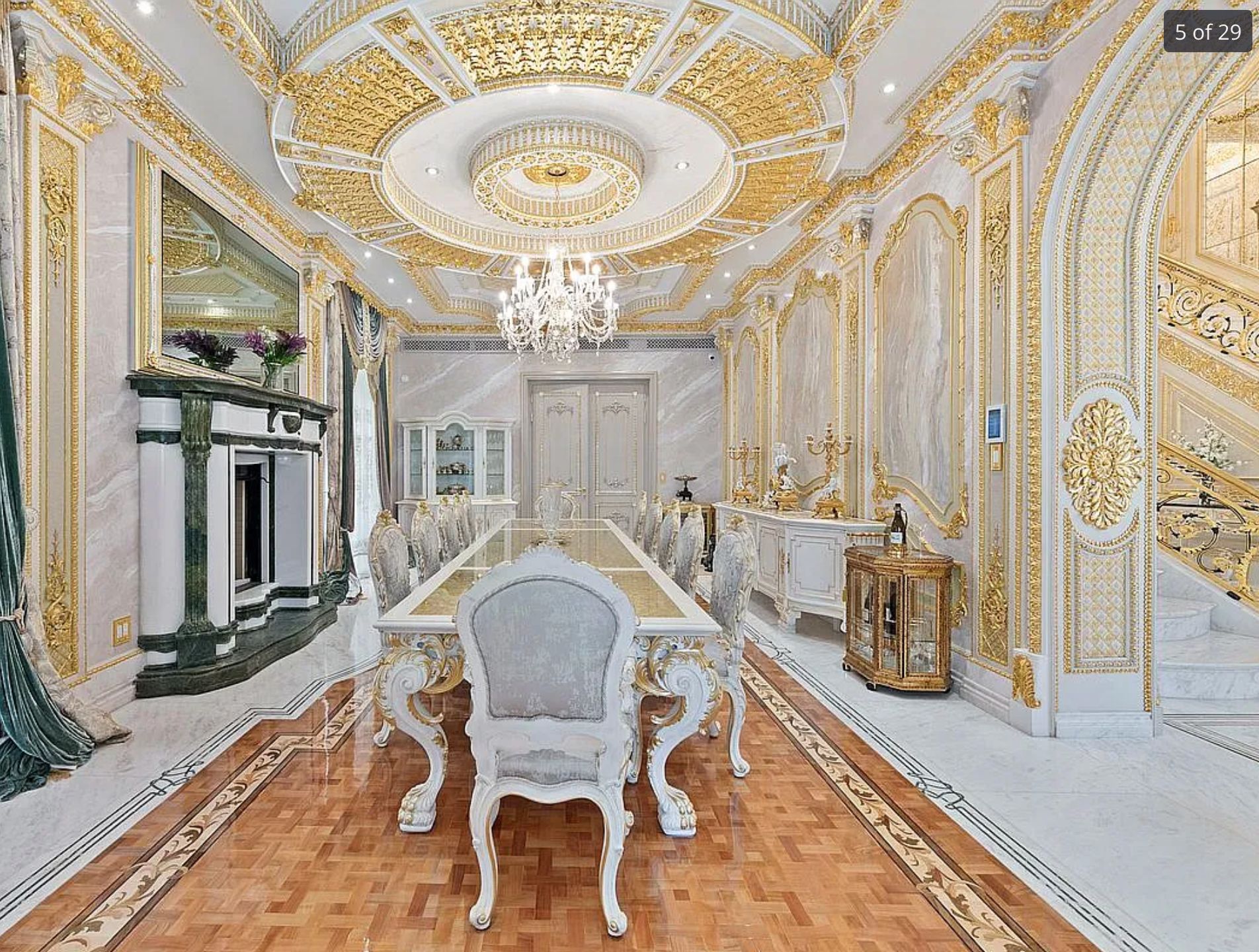 A dining room showing gold and white ornate walls and a gold and white ornate ceiling with a chandelier. There are parquet floors and an ornate long gold and white dining table. 