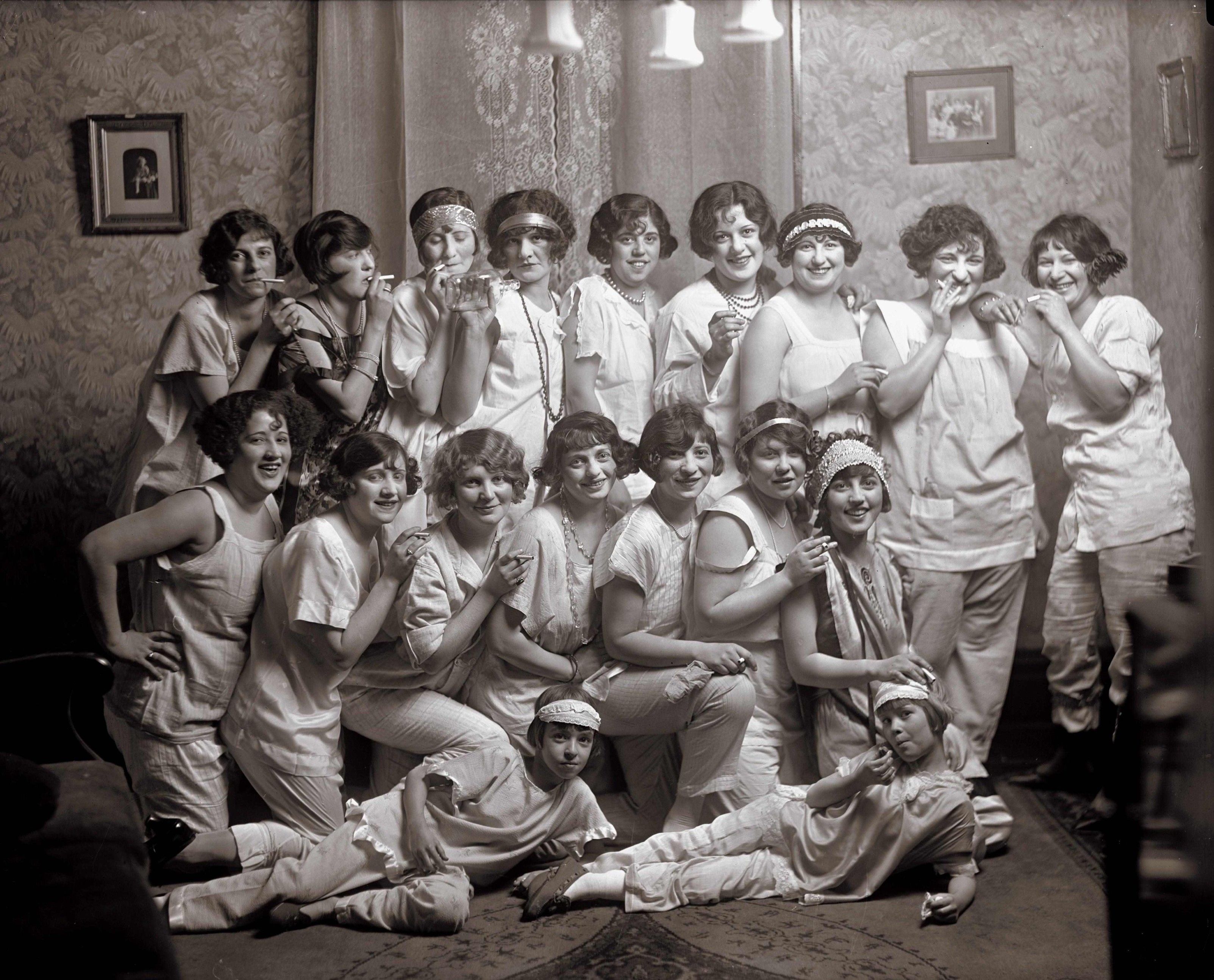 Ein Vintage-Schwarz-Weiß-Foto mit einer großen Gruppe junger Frauen aus den 1920er Jahren, die gemeinsam in einem Raum posieren. Sie sind in historische Kleidung (weiße Schlafanzüge) gekleidet und zeigen eine Mischung aus verspielten und glamourösen Ausdrücken. Einige halten Requisiten wie Zigaretten in der Hand..neenee, es sind Zigaretten!