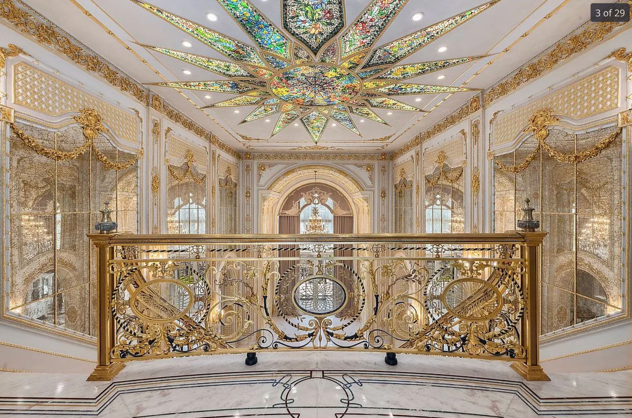 The landing inside an incredibly garish mansion. The floor is marble, the railing is very ornate and gold, the walls are mirrored and gold, there is ornate plasterwork around the ceiling, also gold. There is a rainbow starburst shaped skylight above.