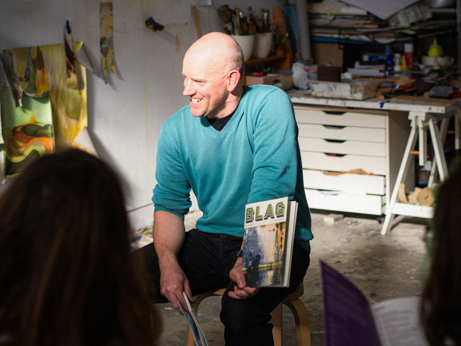 Man sitting on a stool holding a copy of BLAG magazine. He has a shaved head, is wearing a green jumper, and is smiling as he looks at someone to his right. The scene is in an art studio setting, and the man is bathed in sunlight, while other parts of the studio are in the shade.