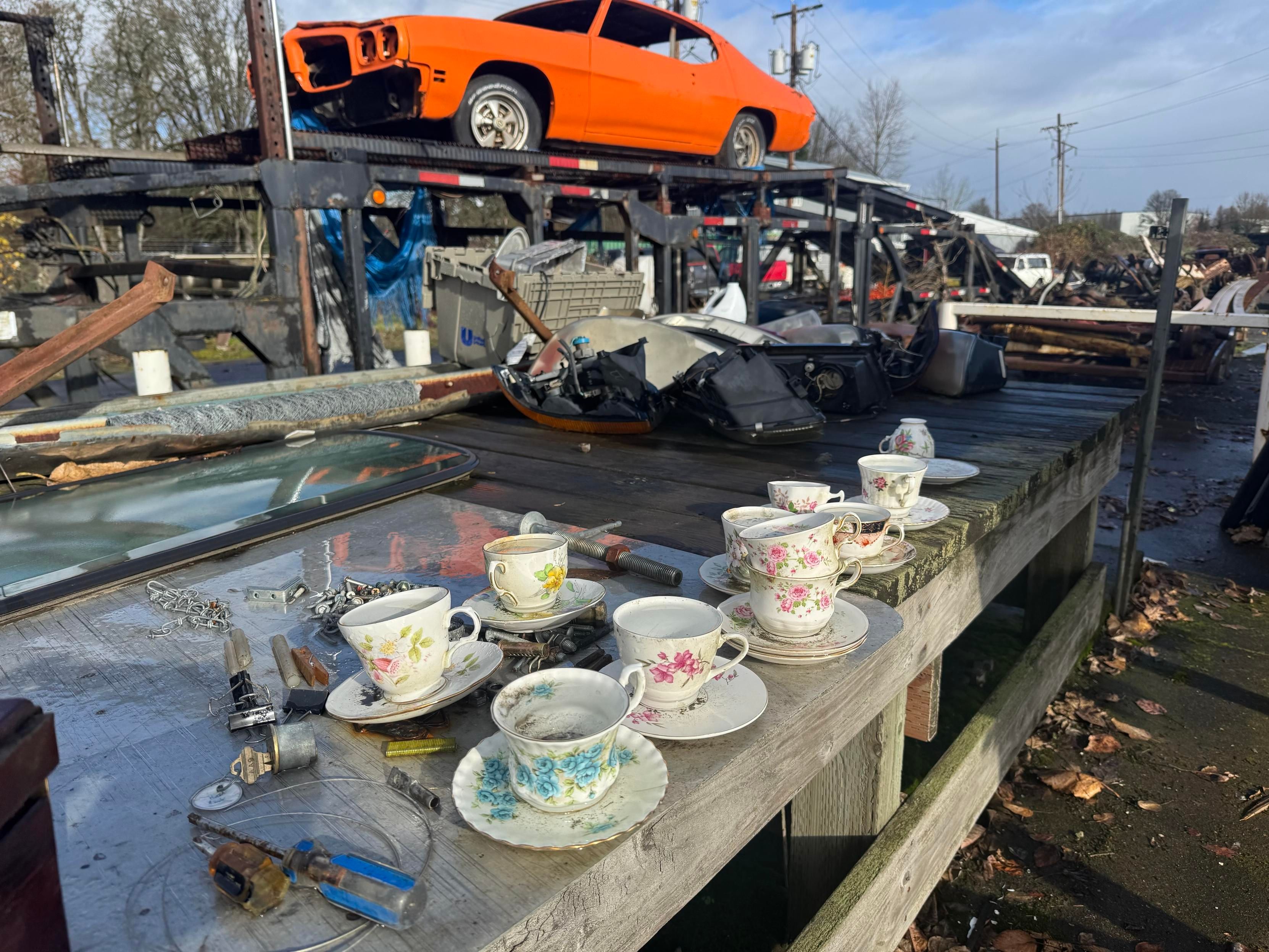 A weathered wooden table displays several delicate floral-patterned teacups and assorted tools, with an orange car partially visible in the background on a trailer. The scene is set in a cluttered junkyard, with various debris and items scattered throughout.