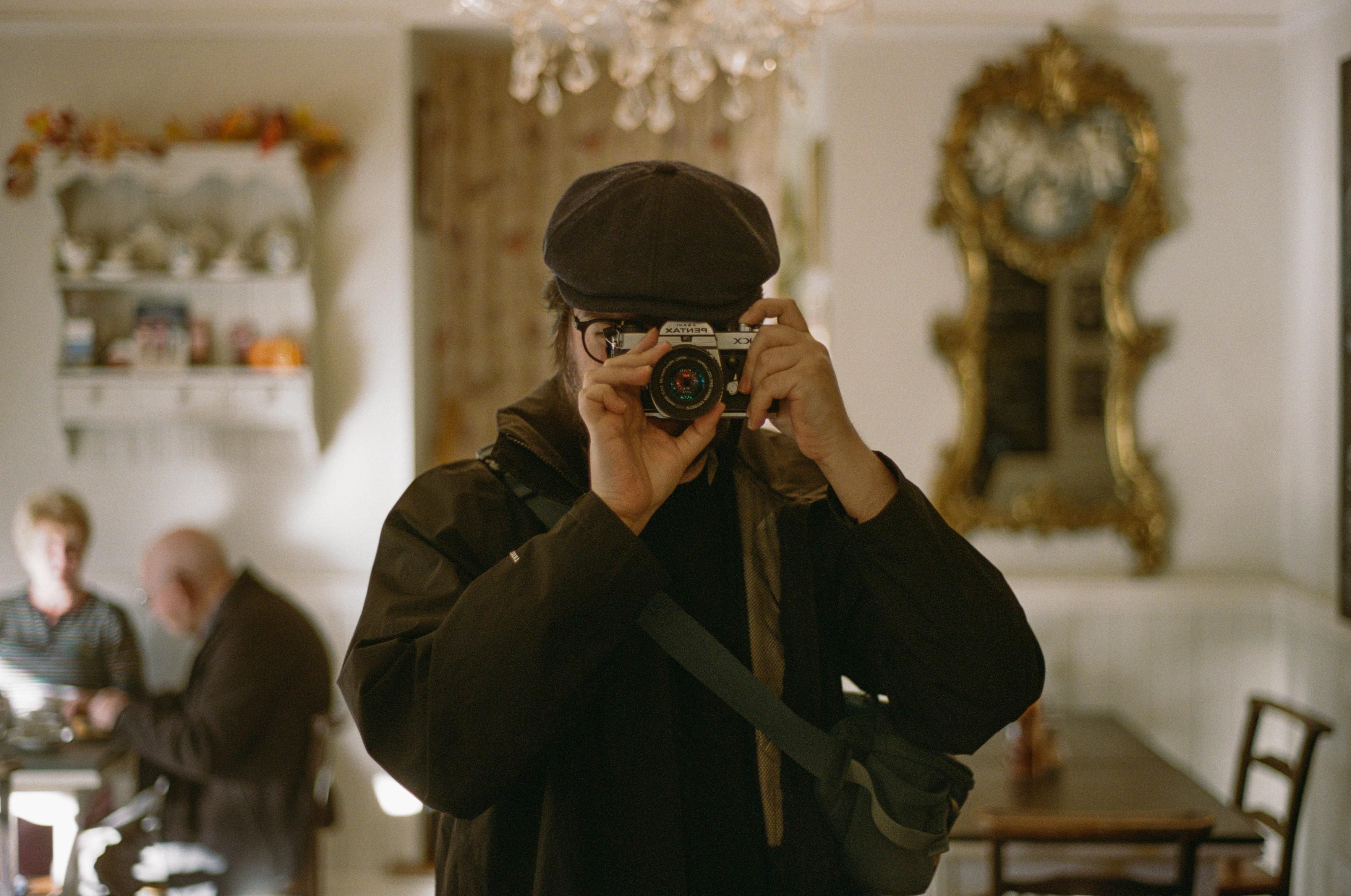A man wearing a flat cap, a black wind jacket and an over the shoulder pouch, holding a Pentax KX camera with both hands in front of his face, reflected in a mirror. In the background, a glass chandelier, antique shelves and wooden tables in the interior of Brady's Tearoom in Grasmere. An elderly couple is sitting at one of the tables.