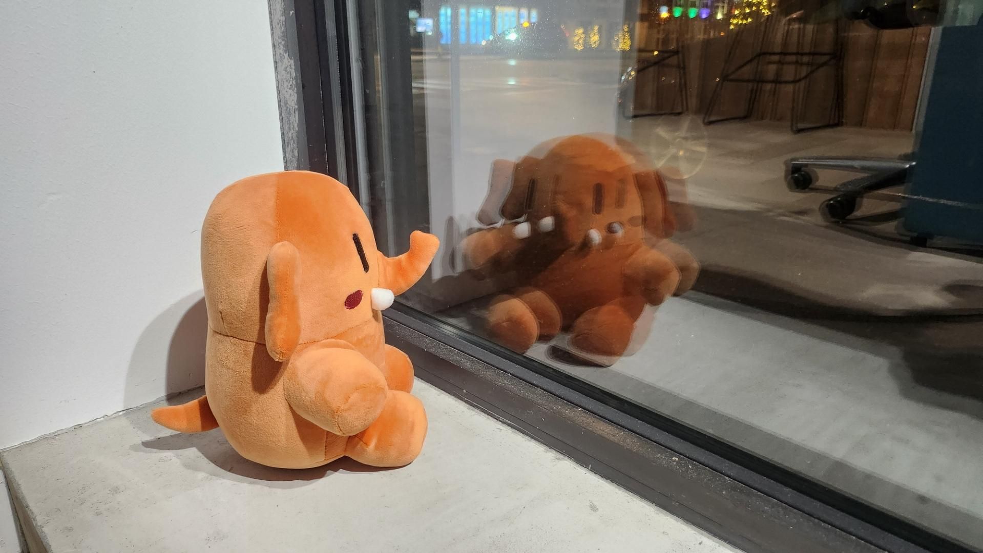 En oransje plushtodon – Mastodons myke maskot – sitter lent mot et vindu og ser ut. I glasset speiler den runde figuren seg tydelig, mens bakgrunnen viser et mørkt uteområde med glitrende lys og kontorstoler innenfor.