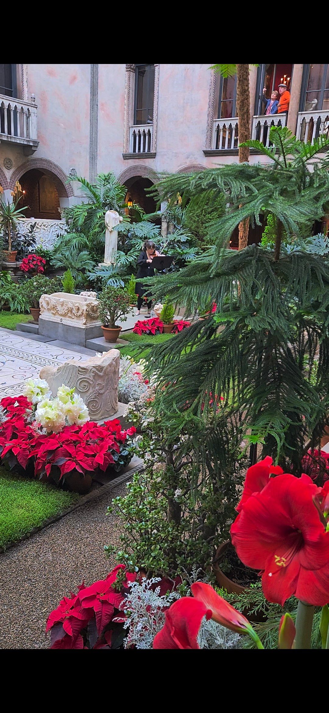 An Italianate courtyard decorated with evergreens, multiple flowering bulbs, and a violinist playing from the middle of the courtyard. 