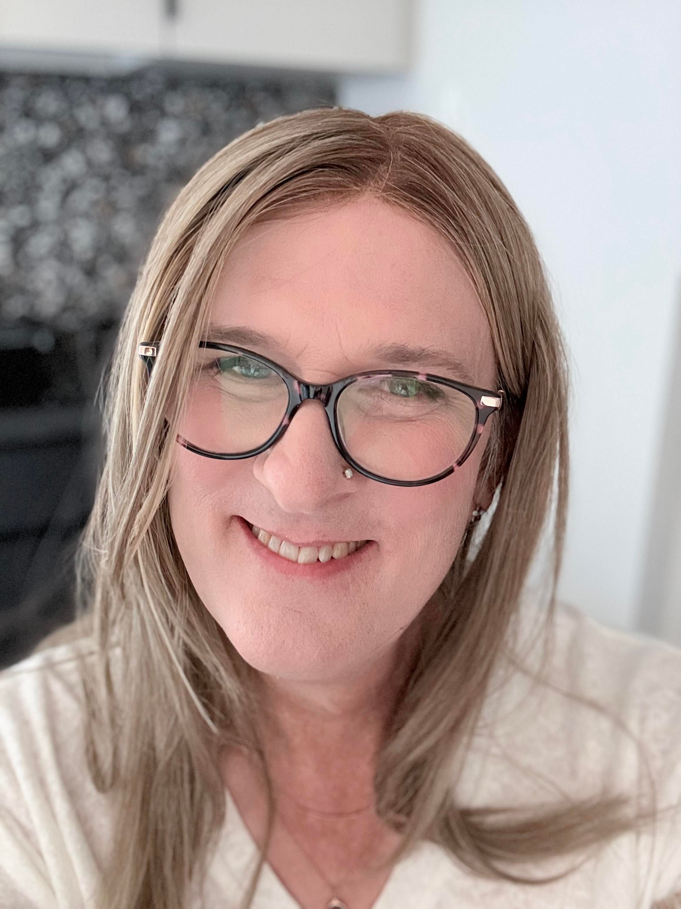 Photo of a smiling Sophie wearing her usual black and pink cat eyed glasses. She has beyond shoulder length blonde hair, is wearing a cream colored v-neck top, and two gold necklaces. She feels pretty!