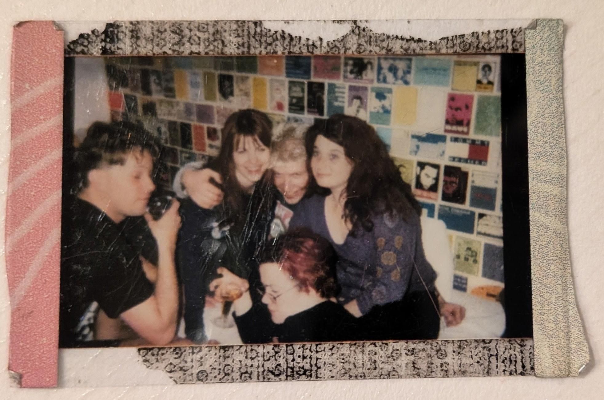 A mini Polaroid with party pink left edge, frayed green right edge, and black top and bottom edges. The photo shows a group of five 20-somethings in a room against a wall of postcards, mostly sitting on a white sofa. From left to right, the top row is the comedy group (as is the unseen photographer): a blond man with shaved sides, wearing a black tee and holding a plastic glass of bourbon; a pretty girl with red hair wearing a black leather jacket; a hipster blond guy with frosted tips wearing a black tee and a gray hoodie; and a beautiful girl with long black hair wearing a blue shirt with gold batik printing. Bottom: a girl with Crayola pink highlights and glasses wearing all black and a look that says she's tired of these loud bitches and all their drama. Which is partly her fault after banging both of the dudes, separately because she's a lady.