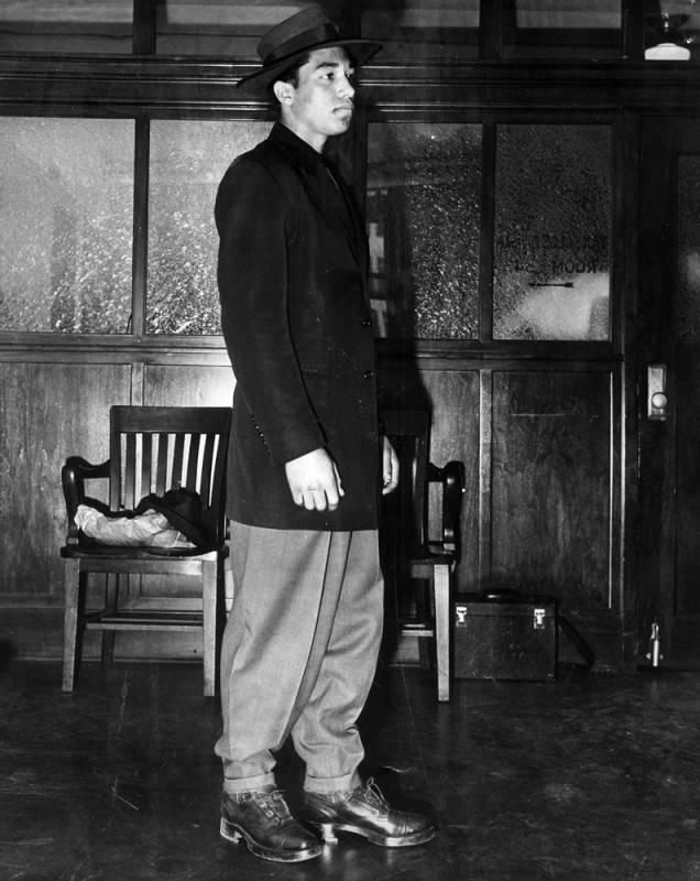 Young main wearing baggy trousers, long coat, and hat.