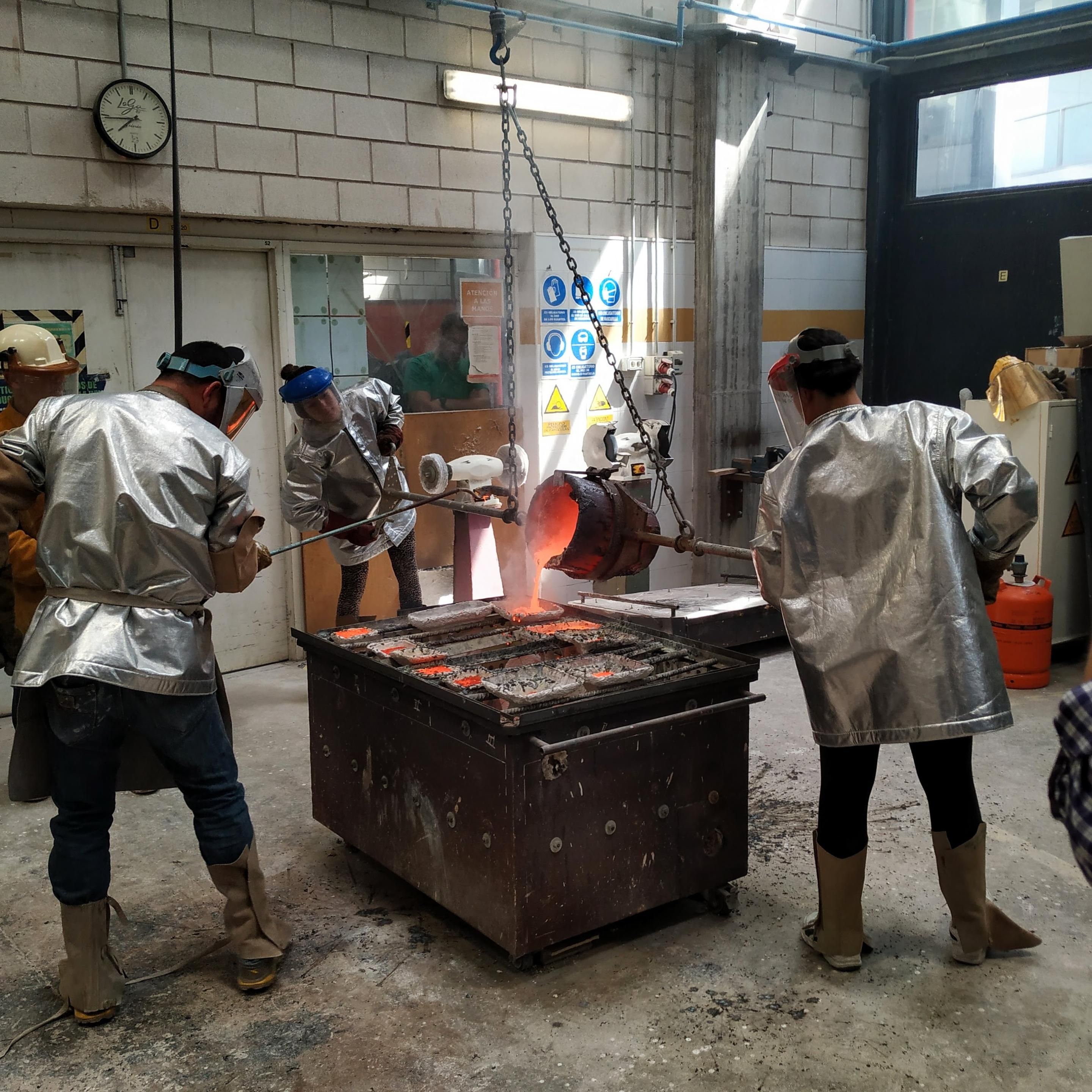 Three people with shiny silver protective clothing and gear, using metal rods to hold a tilted crucible, pouring red hot brass, looking like lava, over a box full of molds for sculptures. 
They're on big, grey classroom/shop lit with natural light coming from windows and skylights.