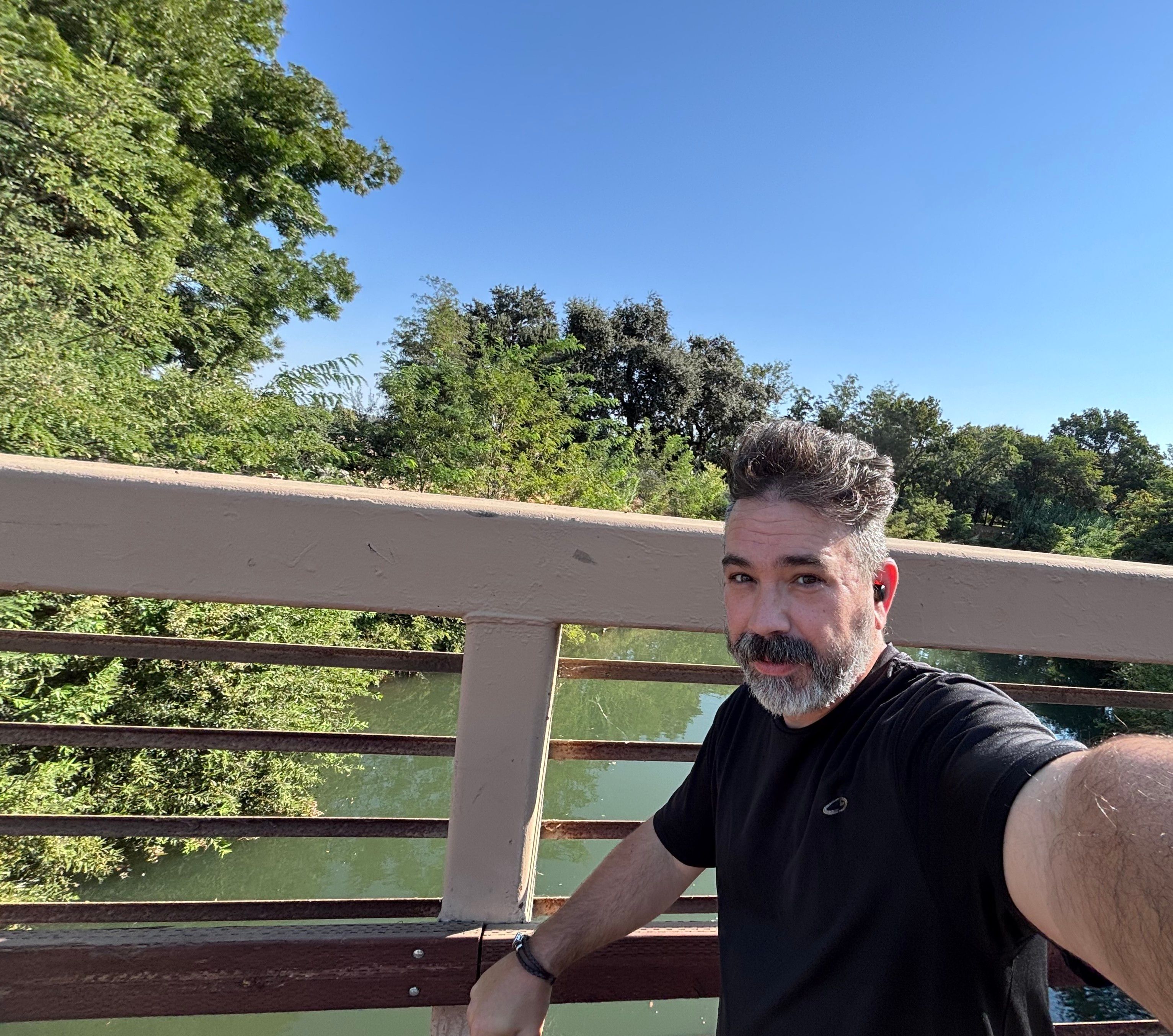 A man with a beard taking a selfie on a bridge over a river, surrounded by green trees under a clear blue sky.